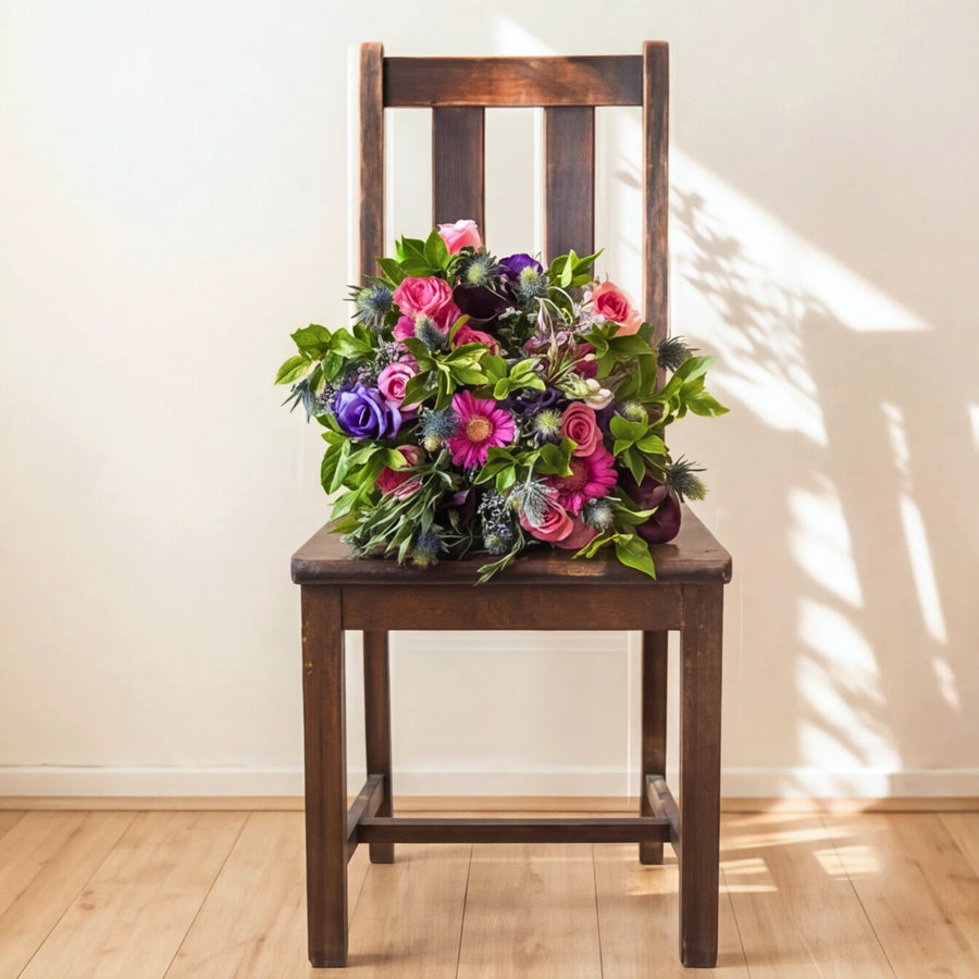 A large, vibrant artificial flower bouquet in shades of pink, purple, and green, displayed on a dark brown wooden chair against a warm beige wall with soft window shadow patterns.
