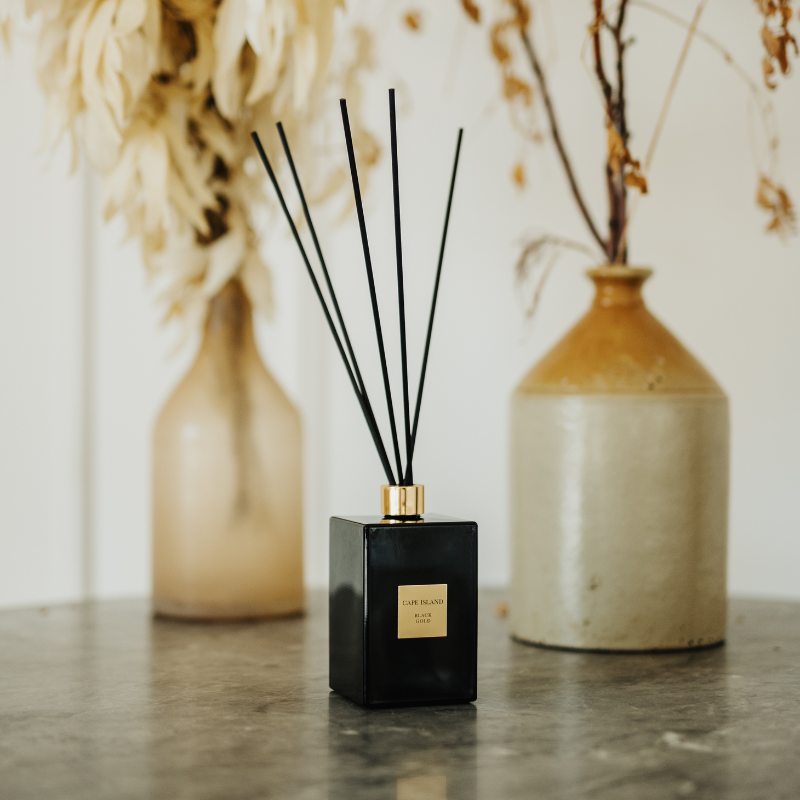 Black reed diffuser with gold cap on a marble surface with decorative vases in the background