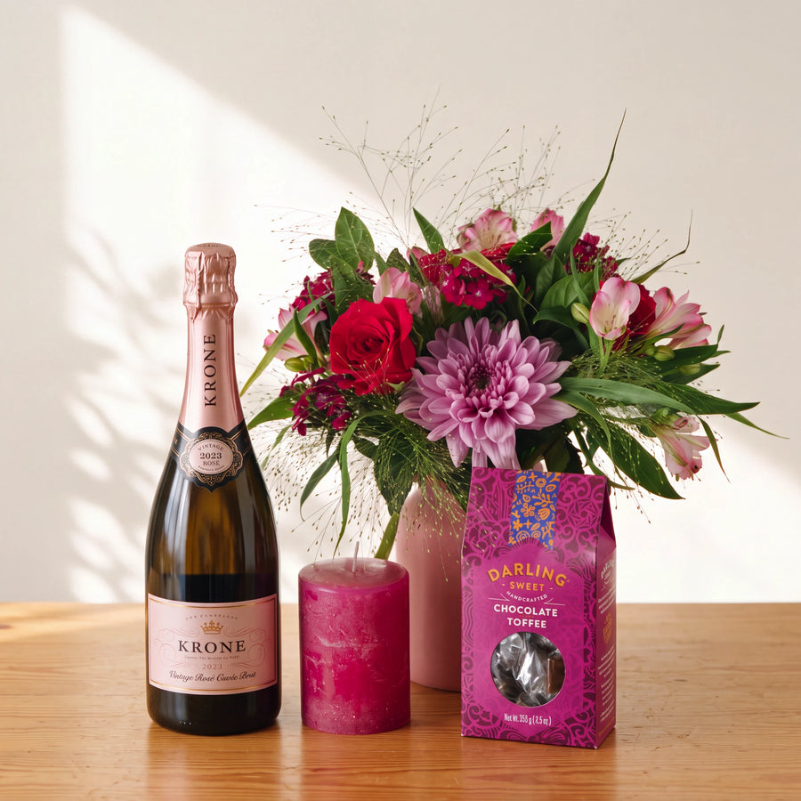 A Krone sparkling wine bottle, a bright pink candle, and a pink box of Darling Cream Chocolate Toffee are arranged on a wooden table beside a pink vase filled with a colorful flower arrangement against a warm off-white wall.
