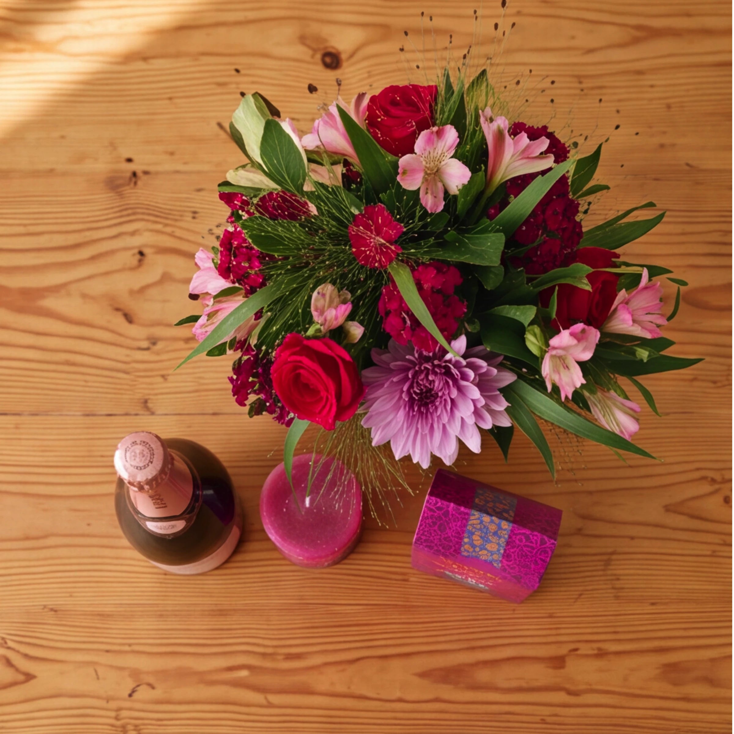 Overhead view of the Bloom and Bubble gift set on a wooden table, a red rose and lilac chrysanthemum jar arrangement beside Krone Rosé Cuvée Brut, a berry-pink candle and Darling Sweet toffee