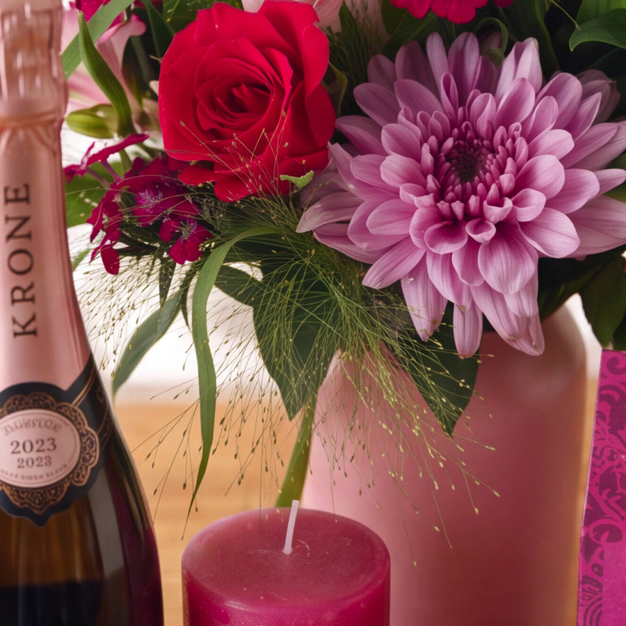 Close-up of the Bloom and Bubble jar arrangement, a red rose and pink-lilac chrysanthemum with alstroemeria and magenta Sweet William beside a Krone Rosé bottle and candle