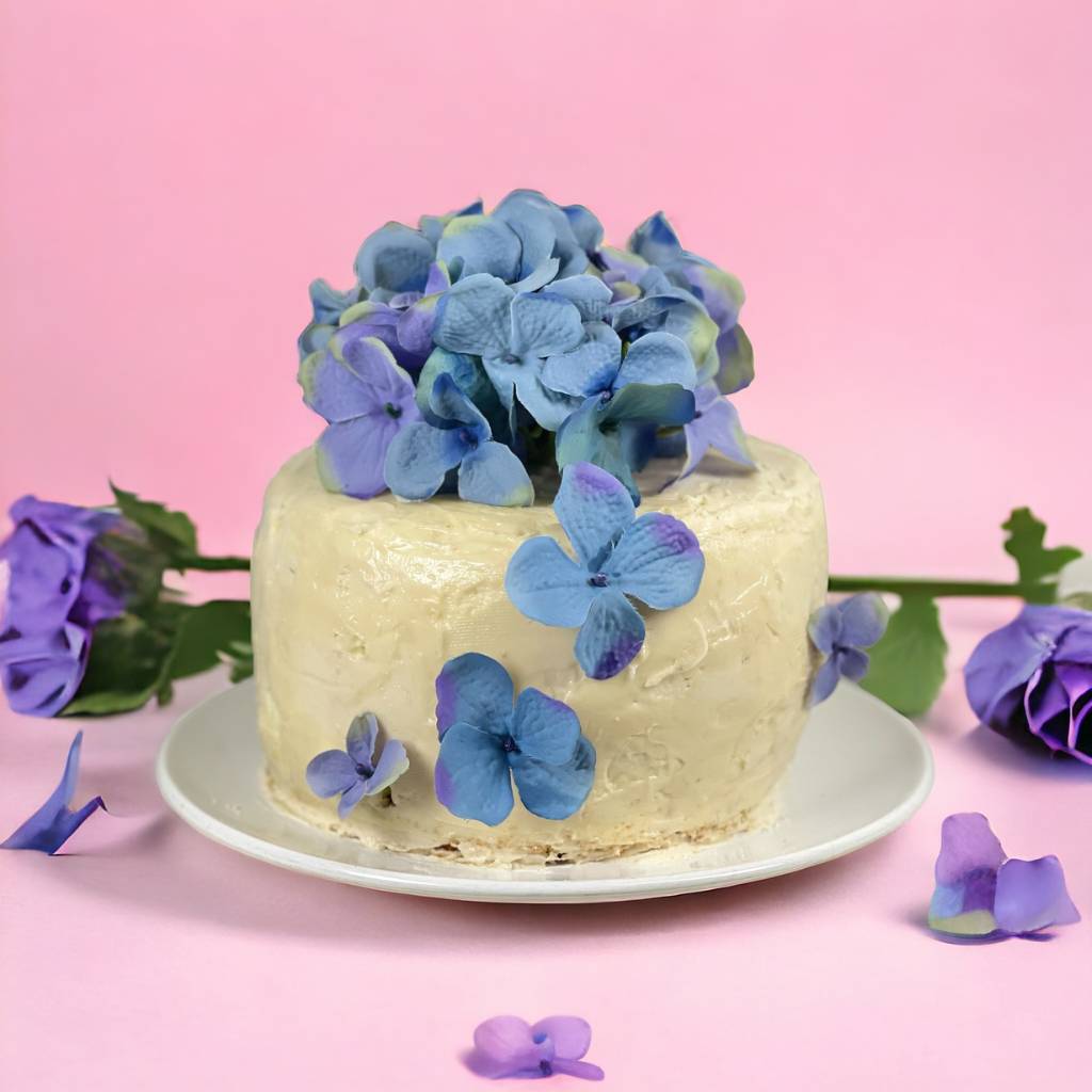 Blue hydrangea topped Madeira cake surrounded by purple flowers by Fabulous Flowers and Gifts.