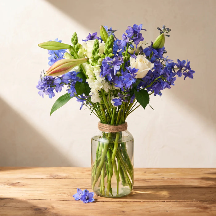 Glass jar vase of blue delphiniums, white roses, snapdragons and lily buds on a wooden table in window light