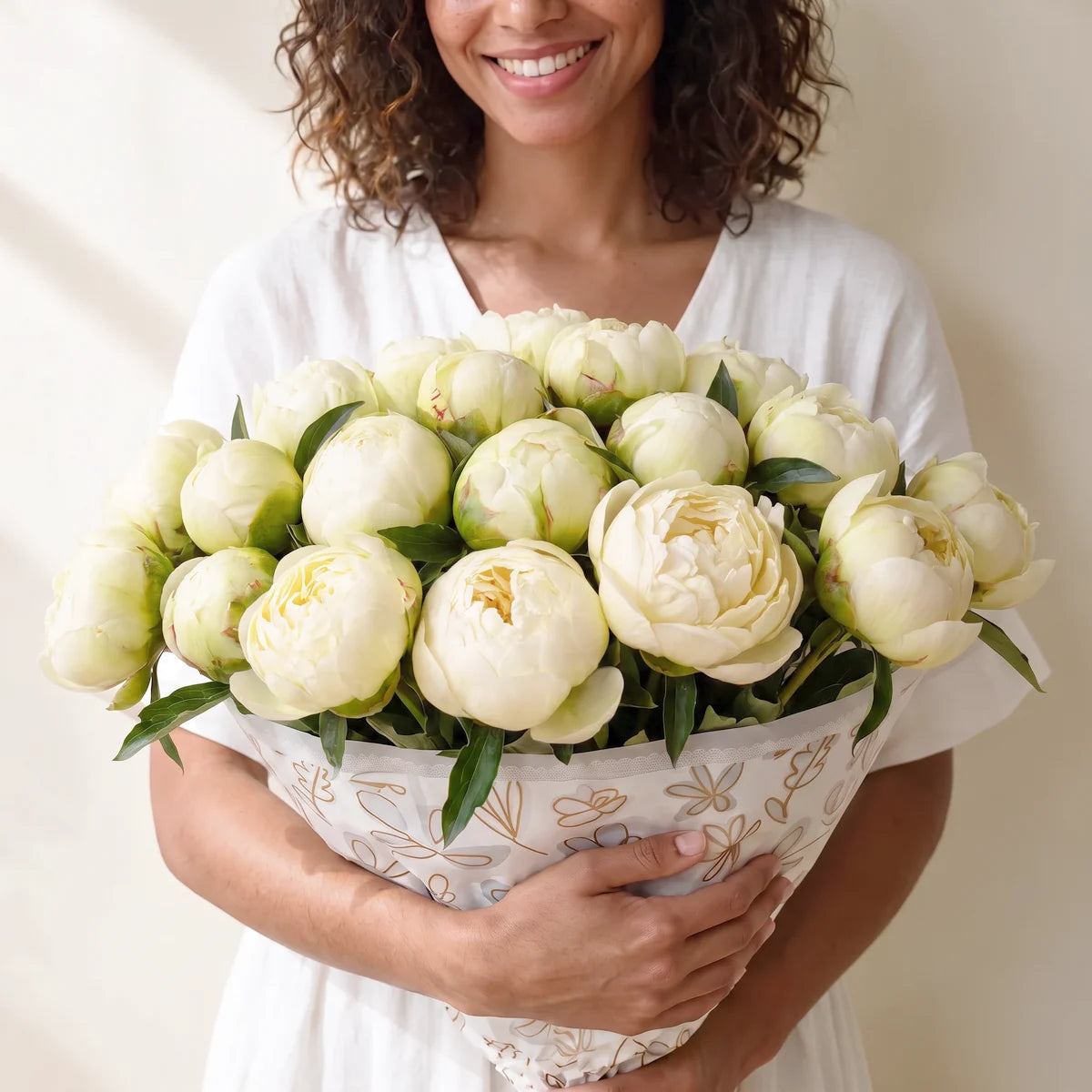 Woman with curly dark hair in a white dress smiling as she holds an armful of twenty white Bowl of Cream peonies, Fabulous Flowers South Africa