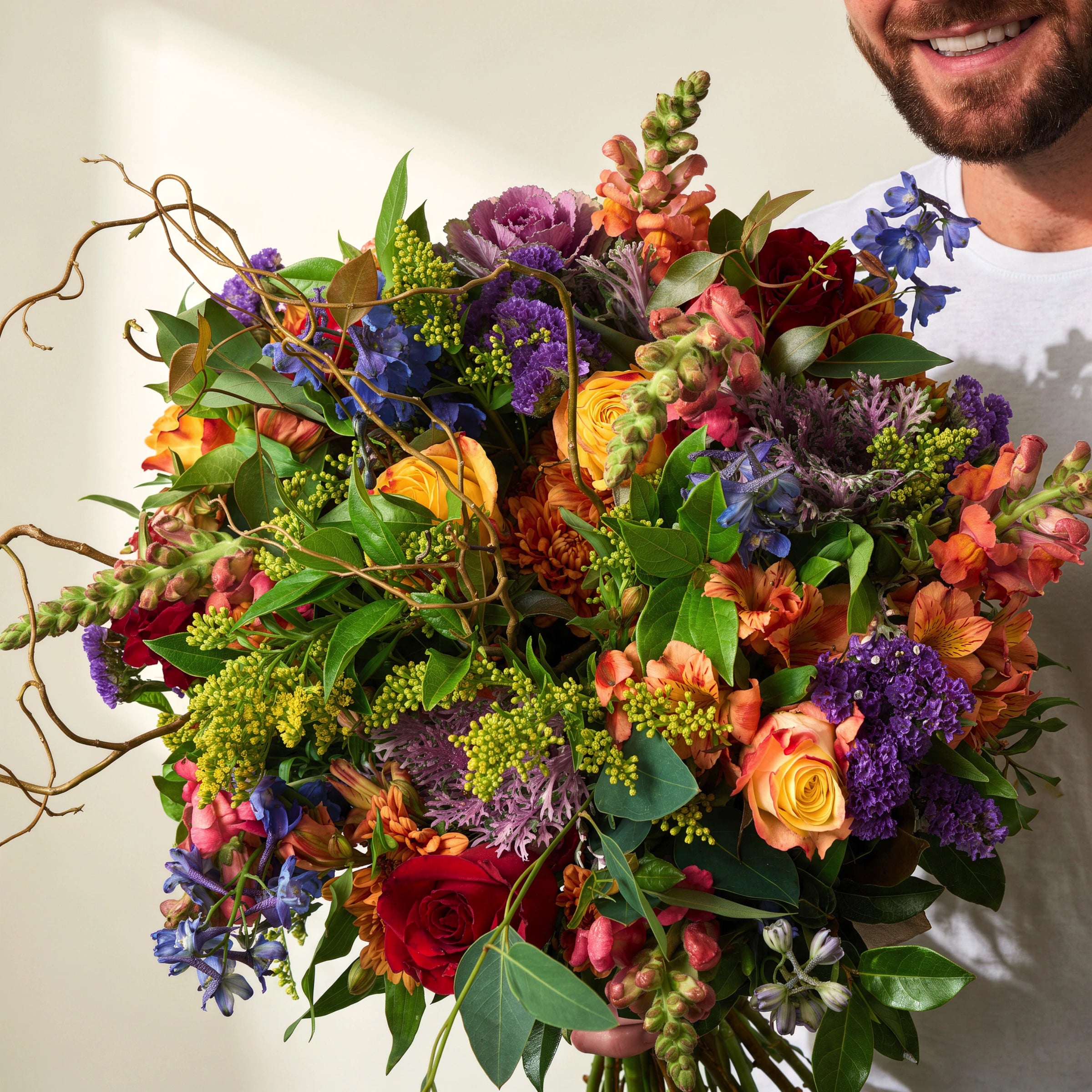 Man in a white t-shirt holding the Brilliantly Happy Bouquet, a vibrant hand-tied arrangement of orange roses, red roses, blue delphinium, purple statice, ornamental kale, snapdragons and twisted curly willow, Fabulous Flowers Cape Town
