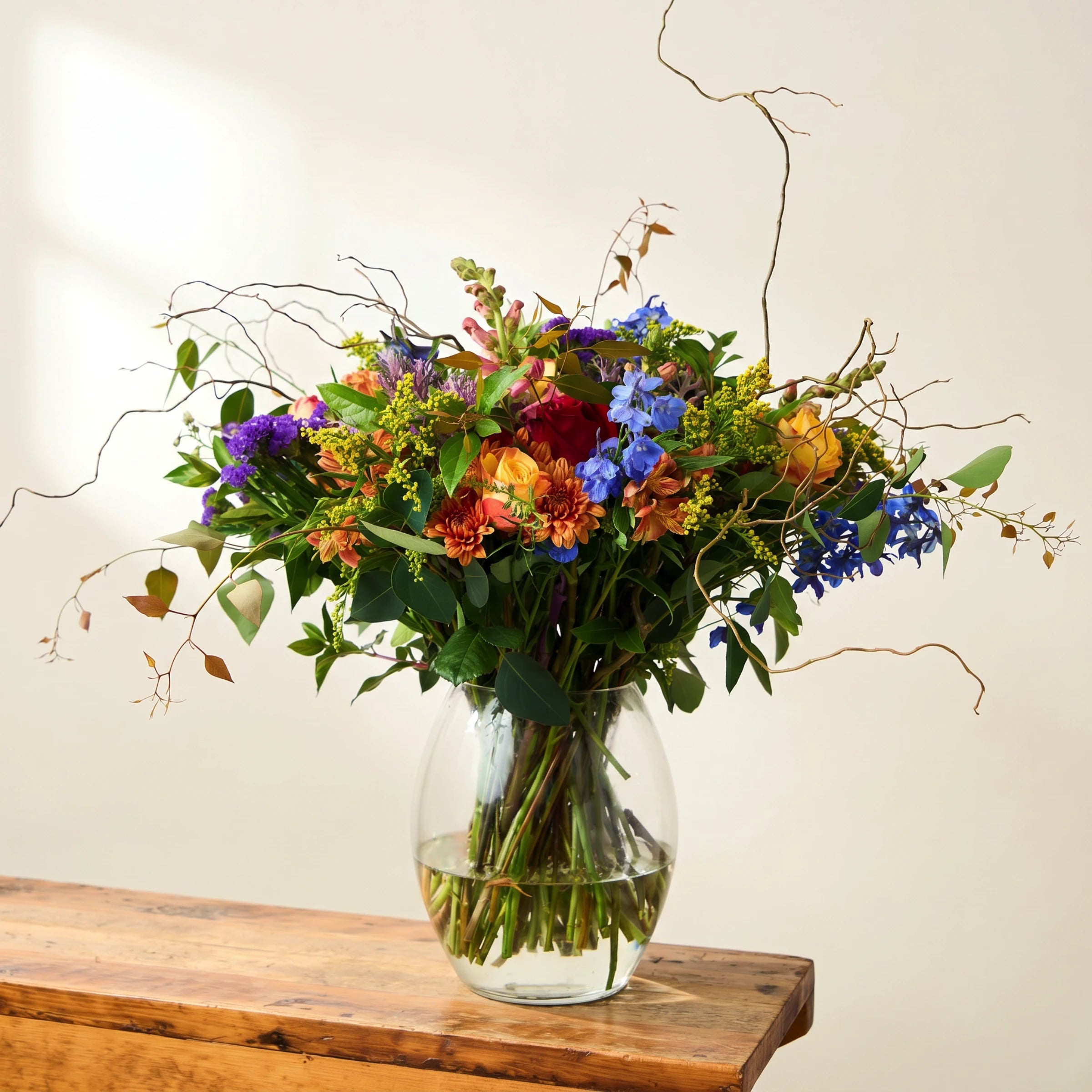 Brilliantly Happy Vase Arrangement on a rustic wooden bench, vibrant mix of orange roses, red rose, blue delphinium, purple statice, orange chrysanthemums, yellow solidago, pink snapdragons and twisted curly willow branches in a clear glass vase, Fabulous Flowers South Africa