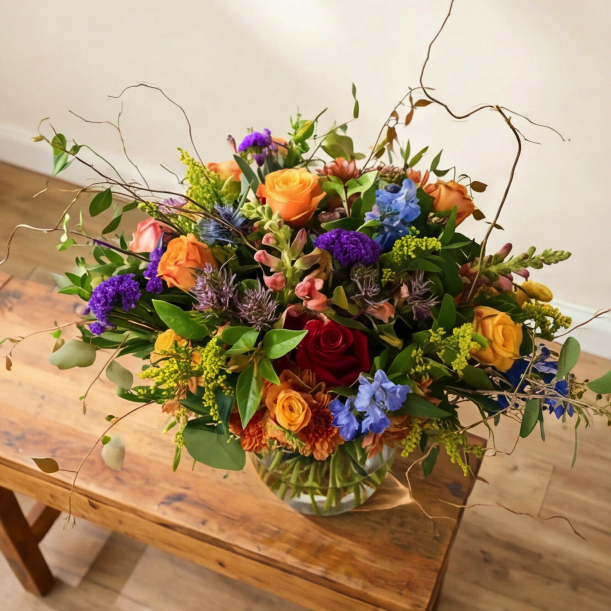 Top-down view of Brilliantly Happy Vase Arrangement showing orange roses, yellow roses, red rose, blue delphinium, purple statice, orange chrysanthemums, sea holly thistle and curly willow in a clear glass vase, locally grown by Fabulous Flowers South Africa