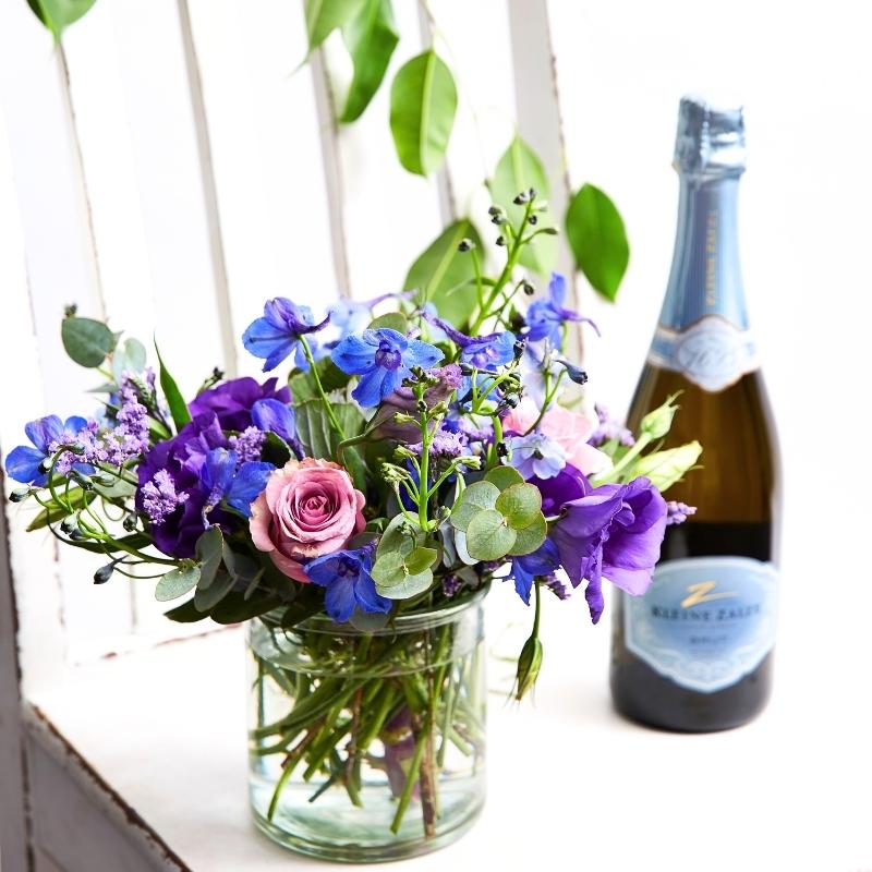 Blue and purple flower arrangement in a glass vase beside a sparkling wine bottle on a bright windowsill