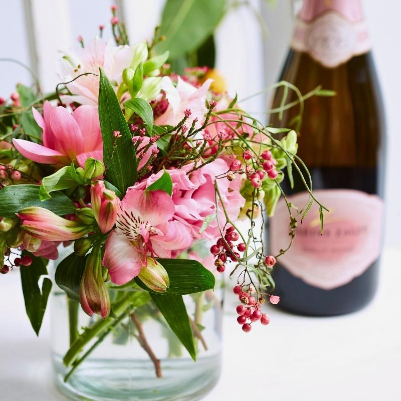 Close-up pink roses and seasonal blooms in a vase with greenery, paired with a bottle of bubbly