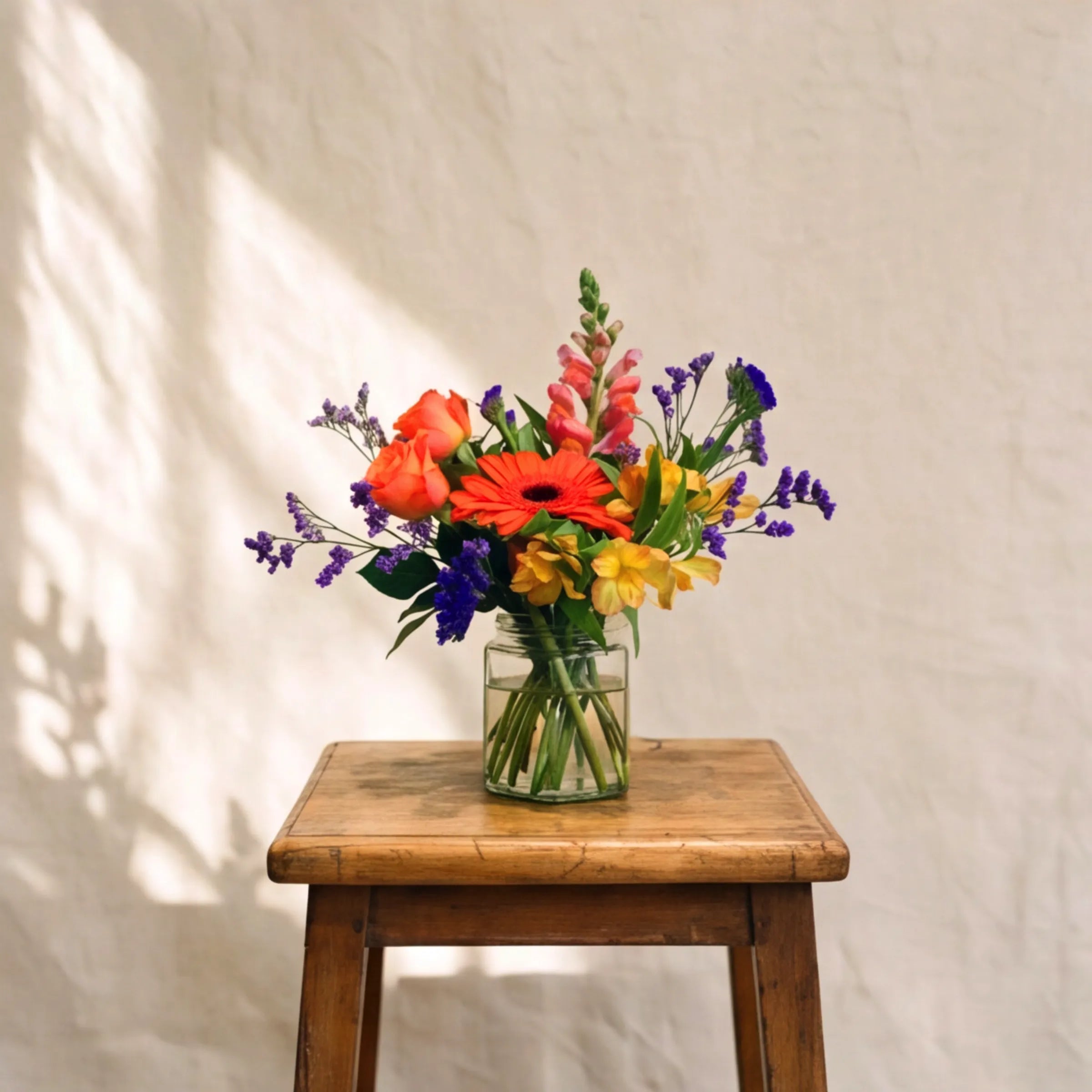 The Carnival in a Jar bouquet by Fabulous Flowers South Africa displayed on a wooden stool, a hand-tied multi-coloured flower posy in a small clear glass jar, ideal as a birthday, get-well or just-because flower gift in Cape Town and Johannesburg