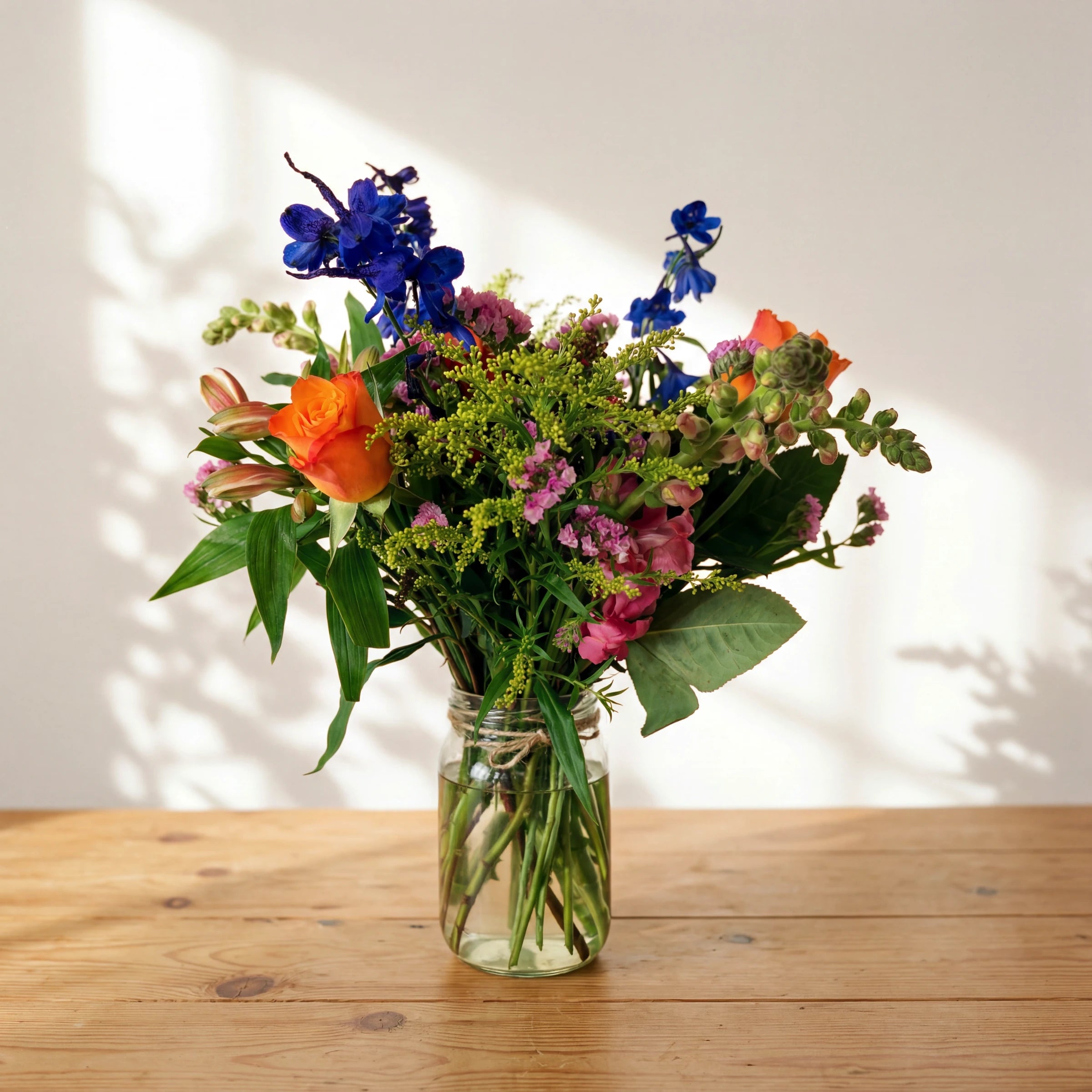 Cheer in a Jar, a bright glass jar of orange roses, blue delphinium and pink limonium on a wooden table, Fabulous Flowers