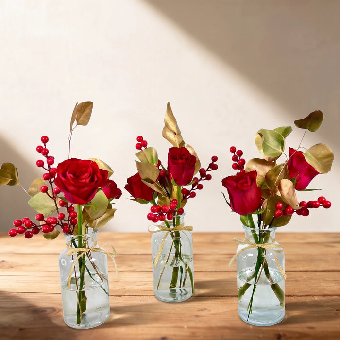 Three small glass bottles of red roses, red berries and gold leaves tied with gold ribbon on a pine table, Fabulous Flowers South Africa