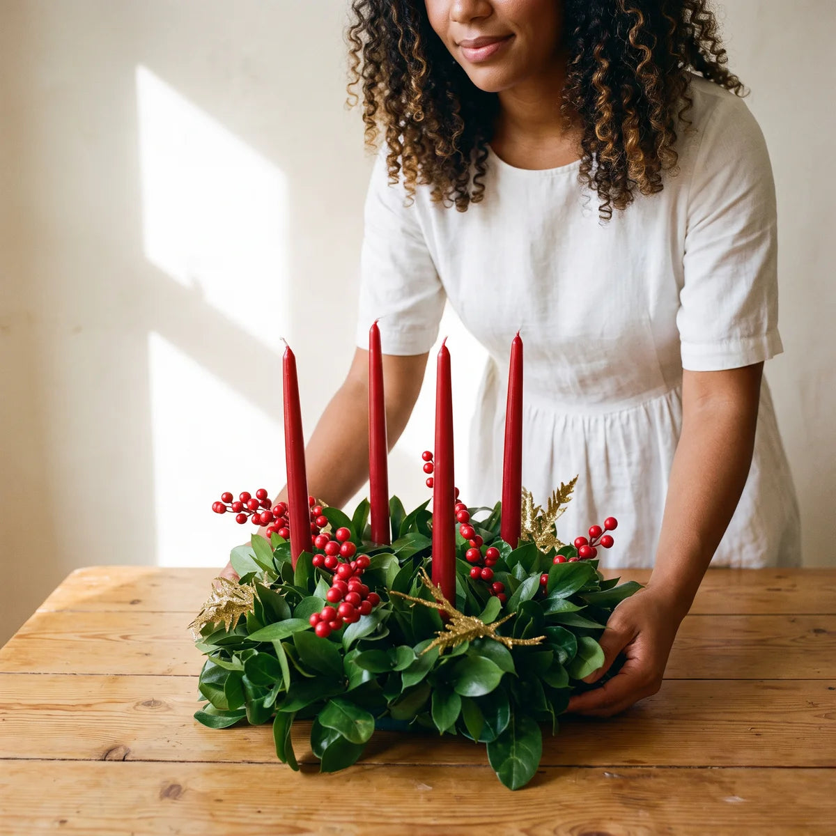 Woman in a white linen dress setting a green wreath with red candles and red berries on a wooden table, Fabulous Flowers South Africa