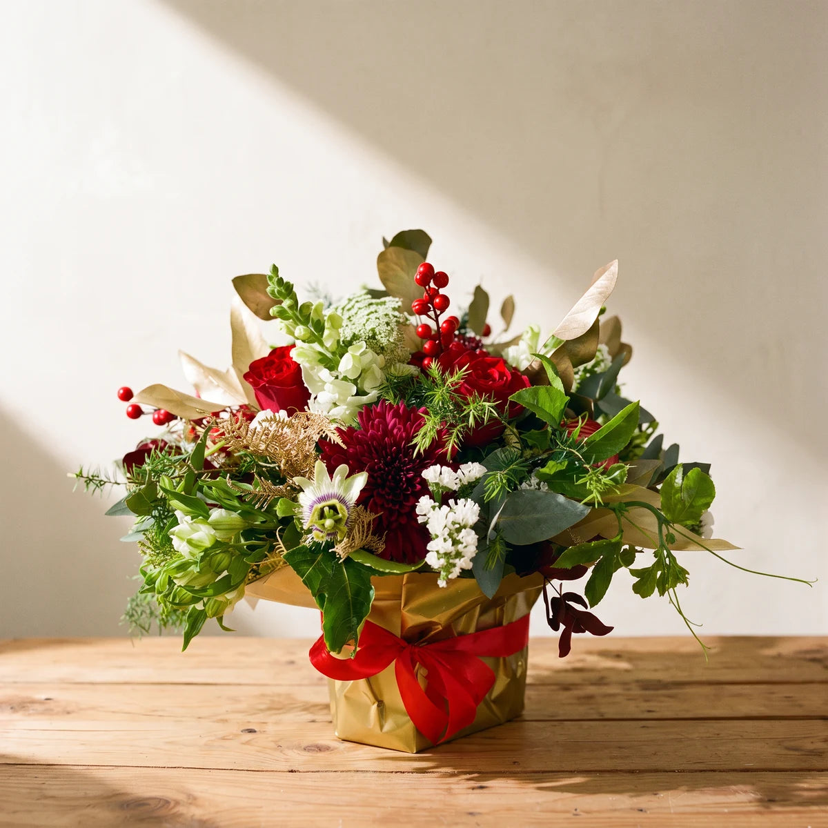 Red and gold Christmas flower arrangement with red roses, white statice and red berries in gold paper on a pine table, Fabulous Flowers South Africa