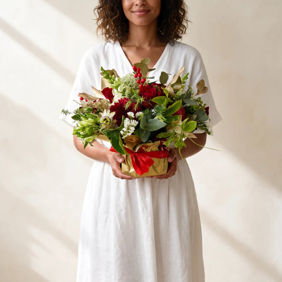 Woman in a white linen dress carrying a red and gold Christmas arrangement with a red bow, Fabulous Flowers South Africa