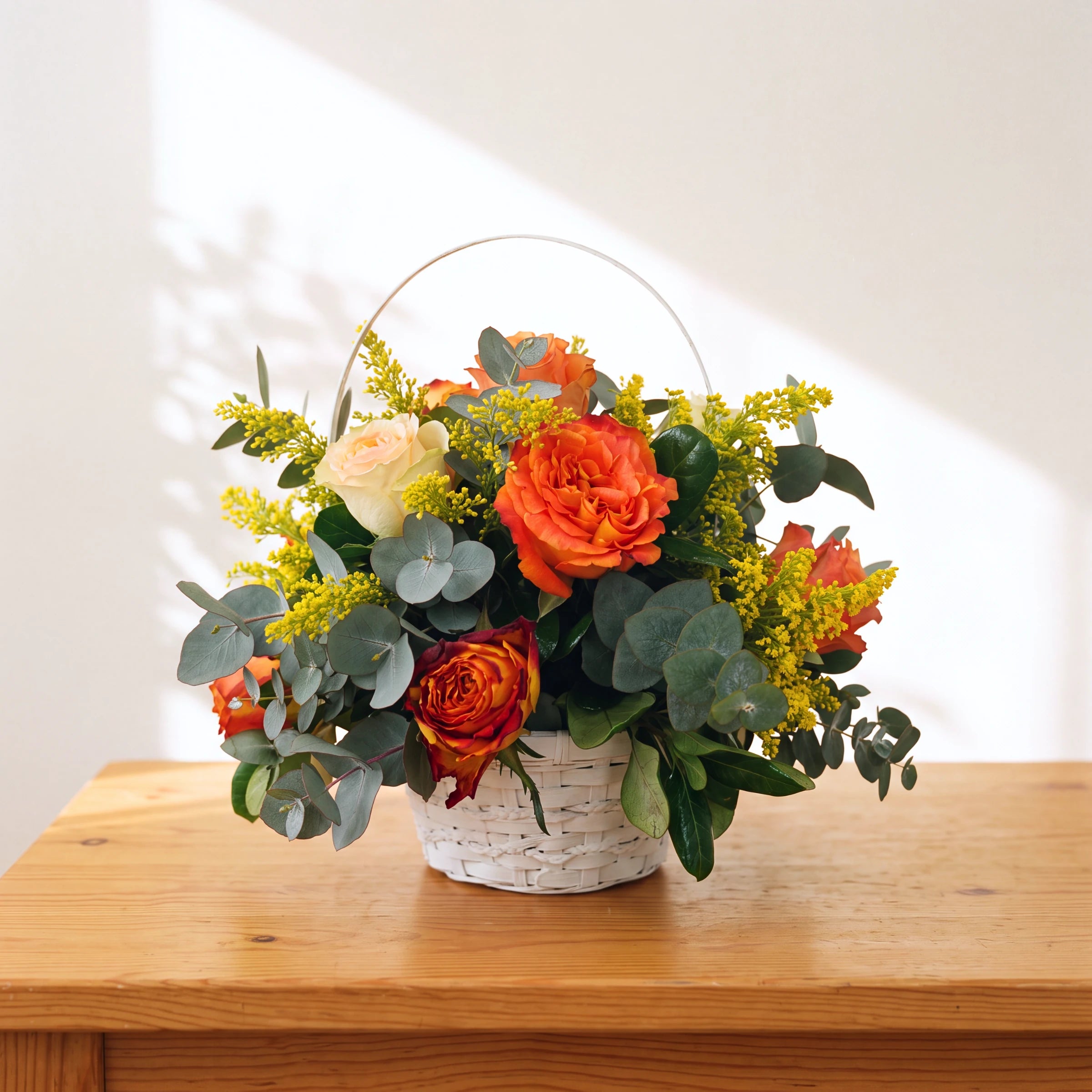 A floral arrangement in a white woven basket with a clear wire handle, placed on a wooden table against a white wall with soft window shadows. The bouquet includes orange roses, green eucalyptus, yellow filler flowers, and a pale peach bloom.