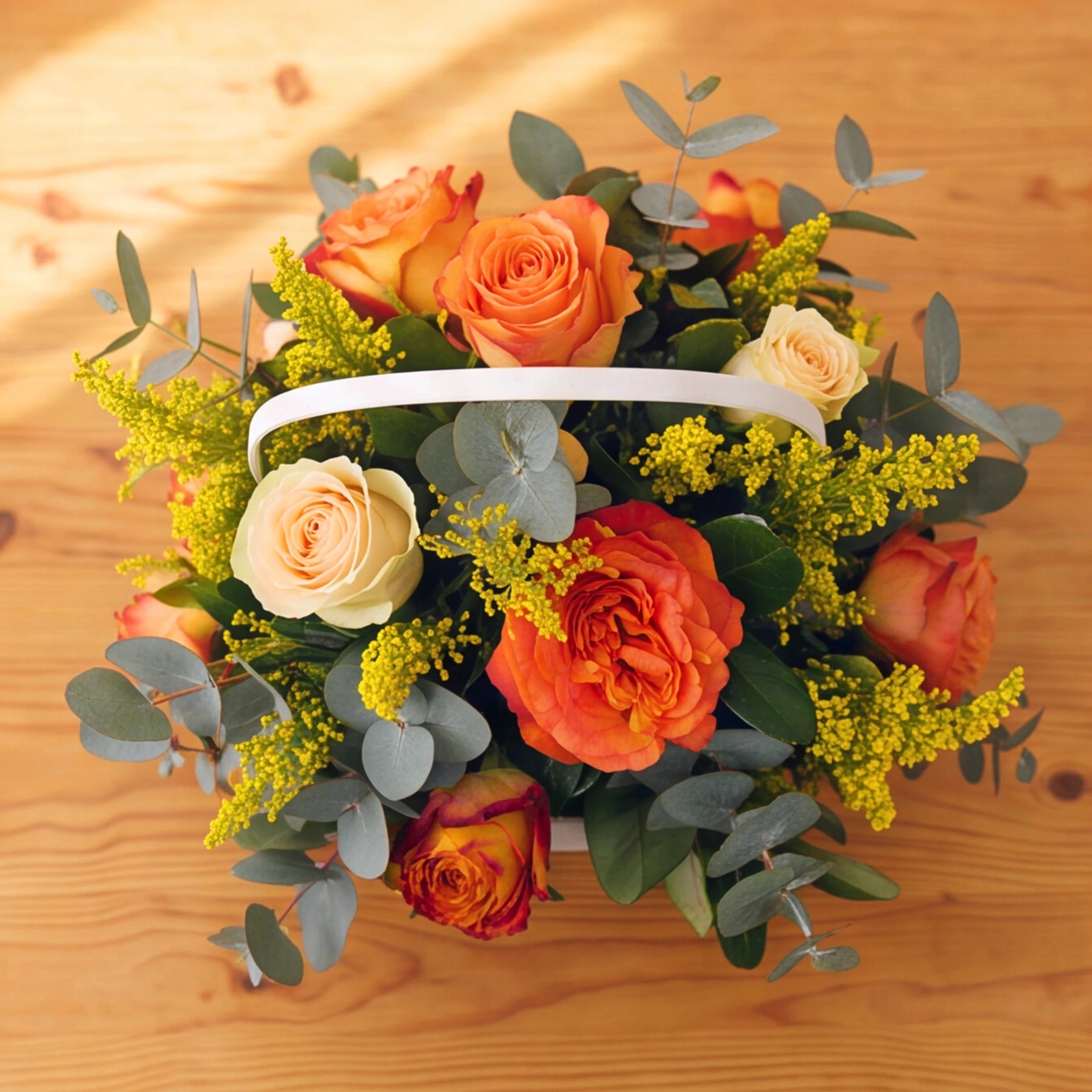 A floral arrangement of orange and peach roses, cream roses, green eucalyptus, and yellow filler flowers in a white basket, placed on a light wood surface.