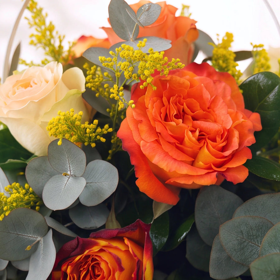 A close-up of a floral arrangement featuring vibrant orange roses, pale peach carnations, red and yellow marigold roses, and dusty blue-green eucalyptus leaves with small yellow filler flowers.