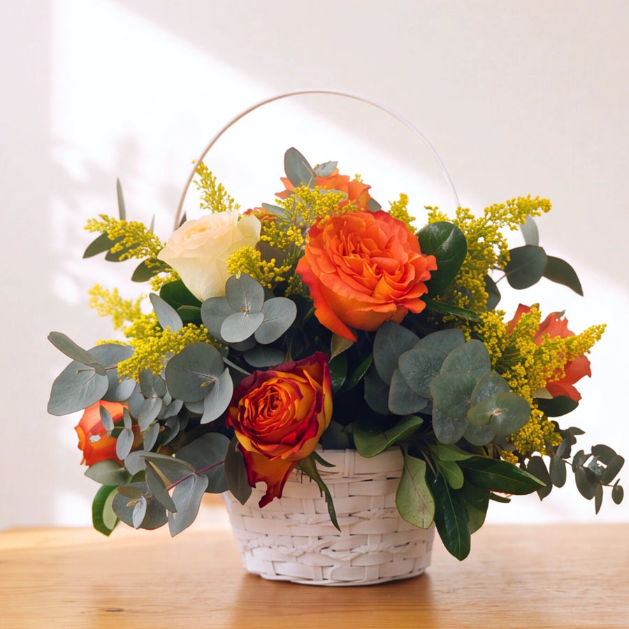 A white woven basket filled with a floral arrangement featuring bright orange roses, green eucalyptus leaves, yellow filler flowers, and a cream rose, set on a light wood surface against a white background with soft window shadows.
