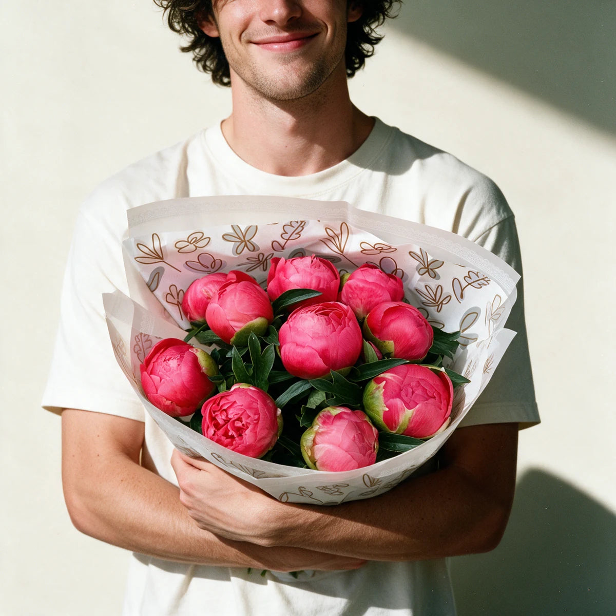 Smiling man in a white T-shirt holding a bouquet of coral peonies against a sunlit wall