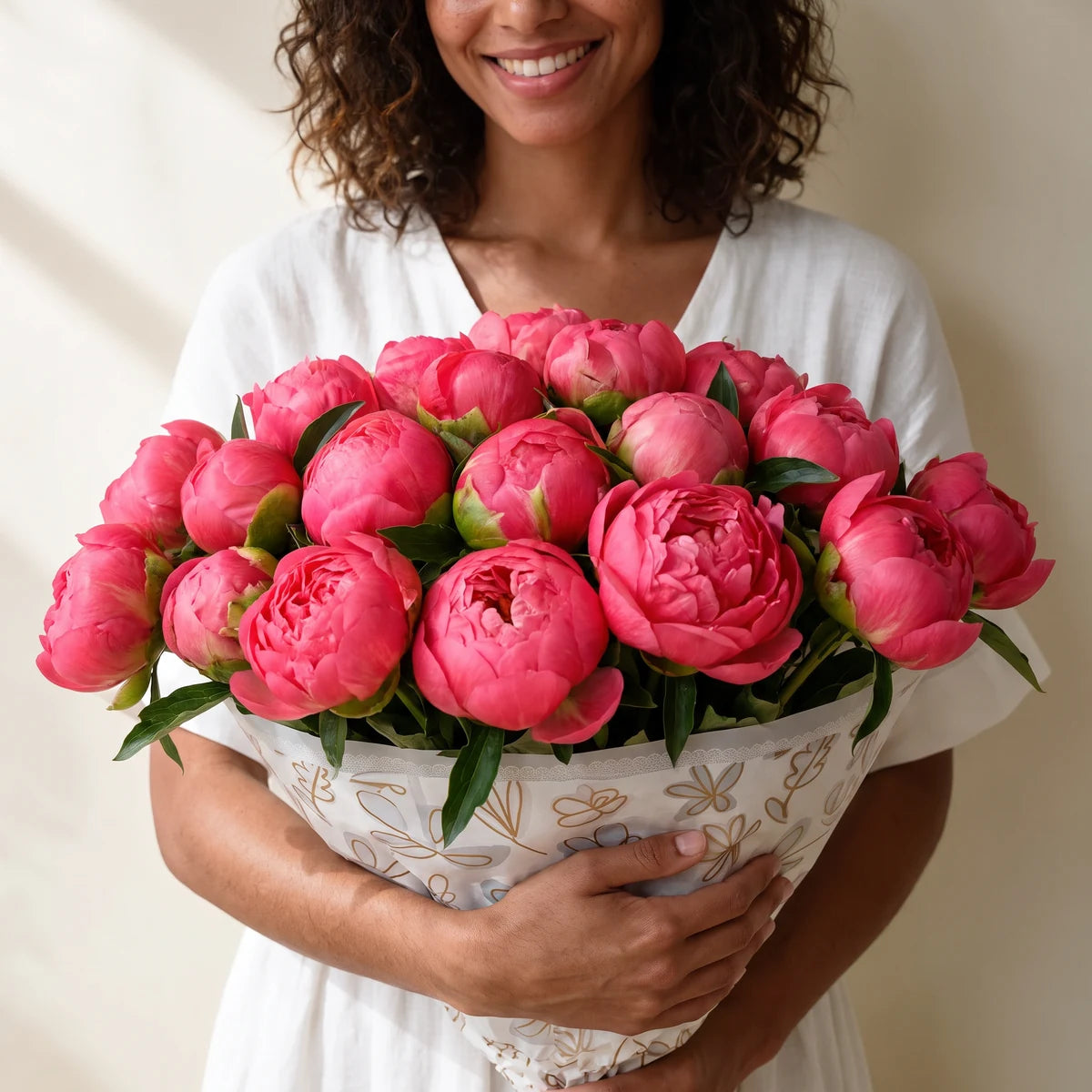 Smiling woman with curly hair in a white dress hugging a large bouquet of coral peonies