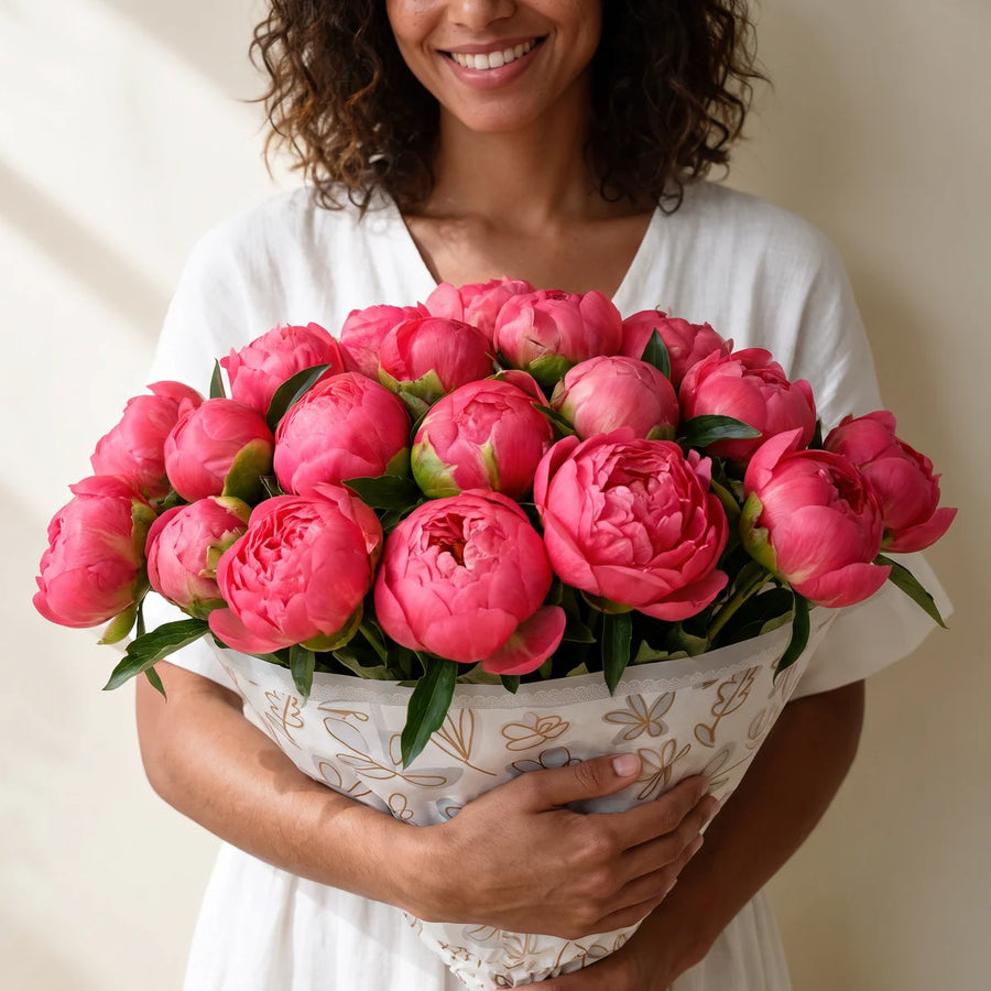 Smiling woman with curly hair in a white dress hugging a large bouquet of coral peonies