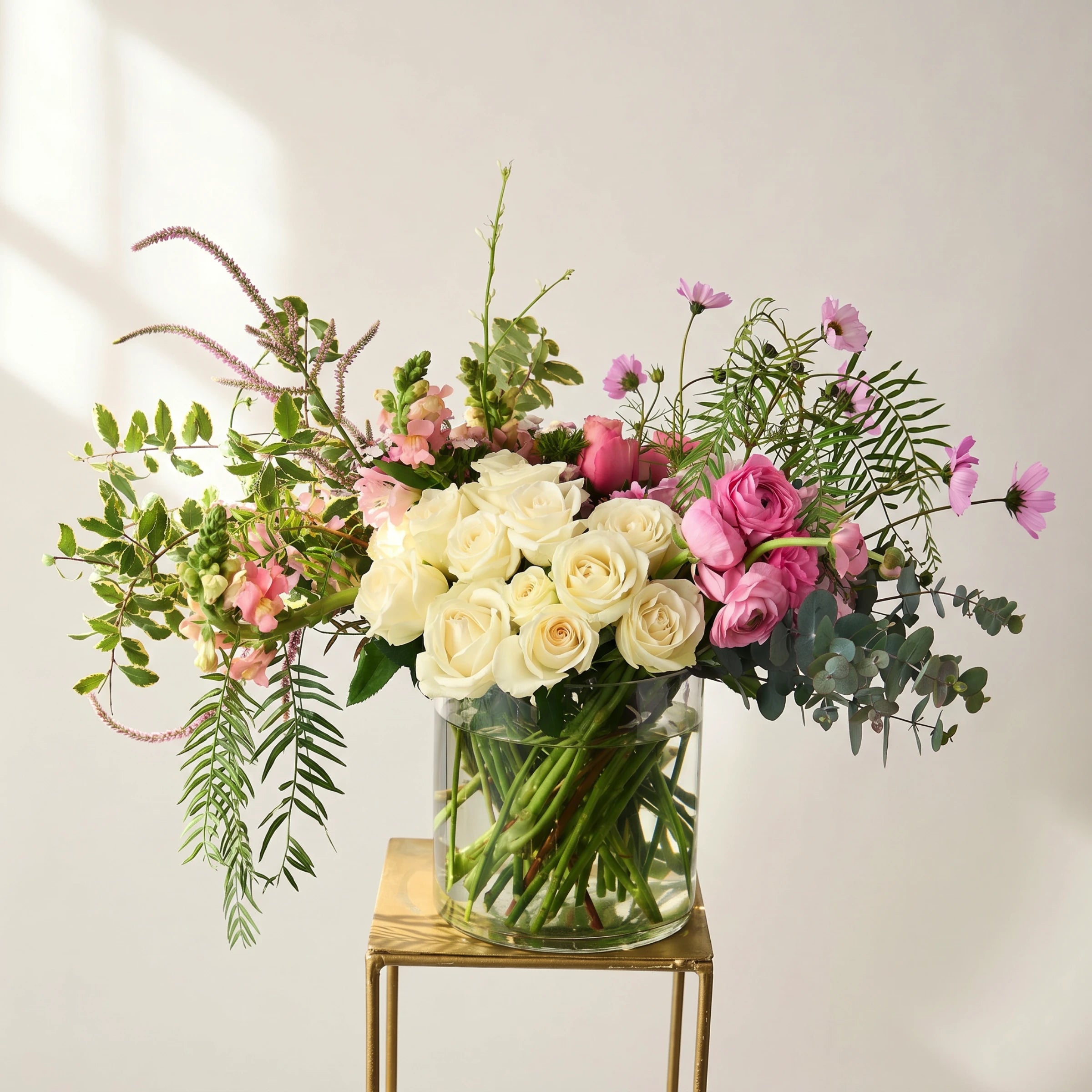 Creative Poetry luxury flower arrangement with cream white roses, hot pink ranunculus, pink cosmos, peach snapdragons, pink veronica and variegated greenery in a clear glass cylinder vase on a gold side table, Fabulous Flowers Cape Town