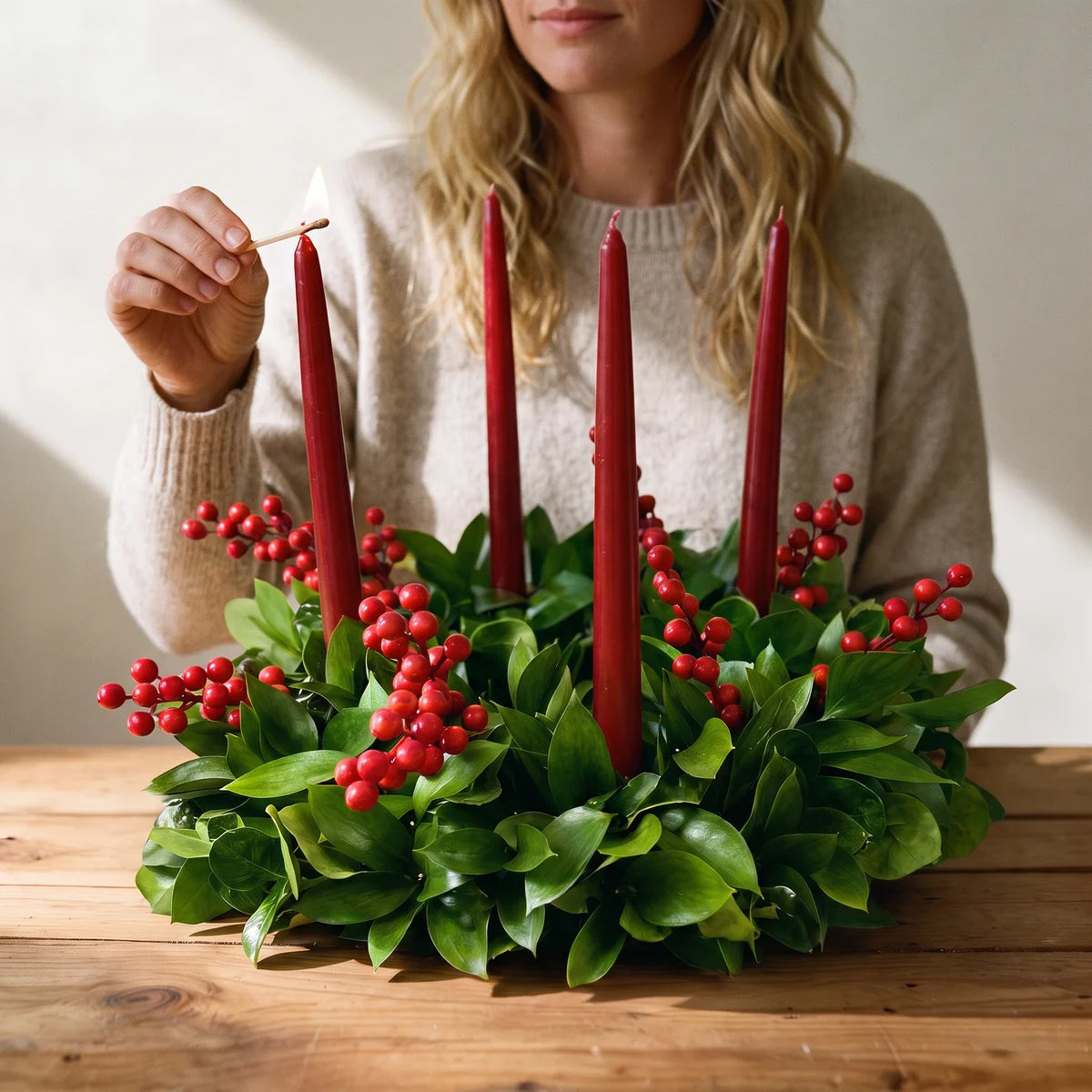 Woman in an oatmeal jumper lighting a red candle on a green wreath with red berries, Fabulous Flowers South Africa