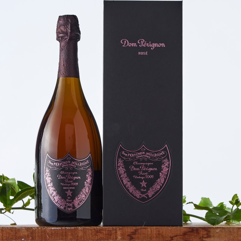 Bottle of Dom Pérignon Rosé Vintage 2009 Champagne with signature pink label, presented beside its black gift box on a wooden shelf with green foliage.