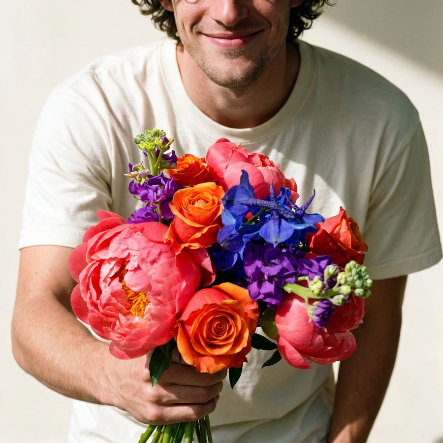 Smiling man in a white T-shirt holding out a bouquet of coral peonies, orange roses, blue delphinium and purple stocks