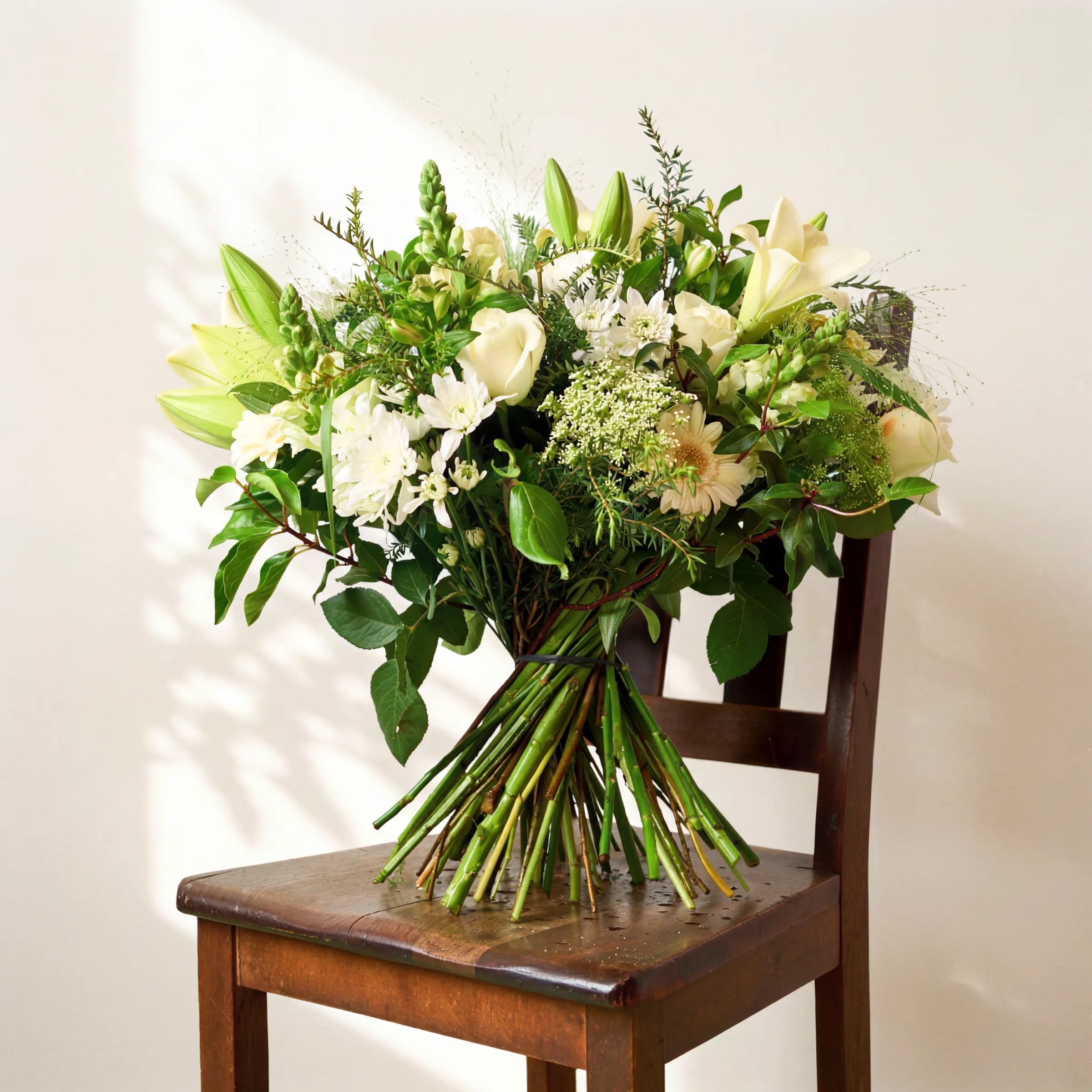 The Elegant White Bouquet hand-tied with white lilies, roses and Queen Anne's lace, resting on a wooden chair