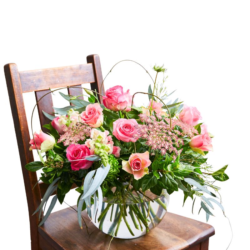 Arrangement of pink roses and greenery in a clear vase on a wooden chair with a white background