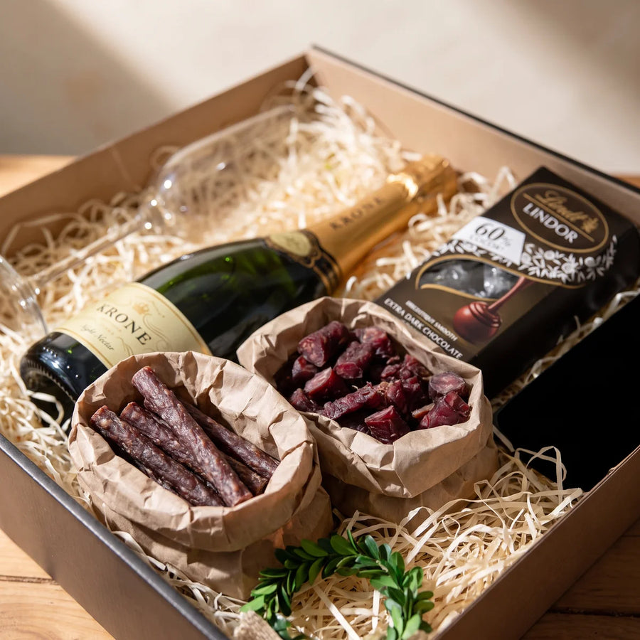 Droe wors and sliced biltong in brown paper bags beside a bottle of sparkling wine and a box of dark chocolate, Fabulous Flowers South Africa