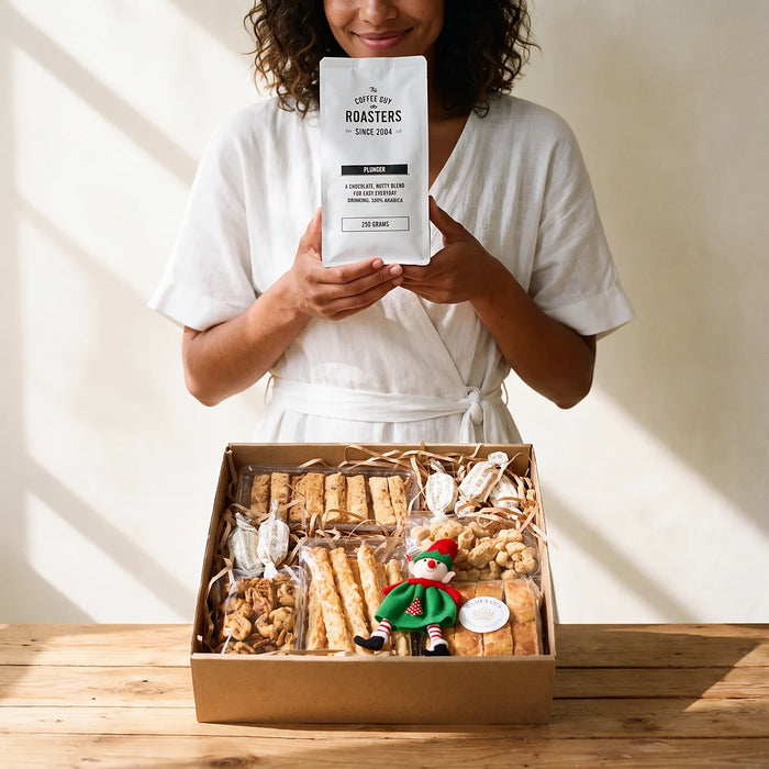 Smiling woman in a white wrap dress holding a bag of The Coffee Guy Roasters plunger coffee above an open Christmas coffee gift box on a pine table, Fabulous Flowers South Africa