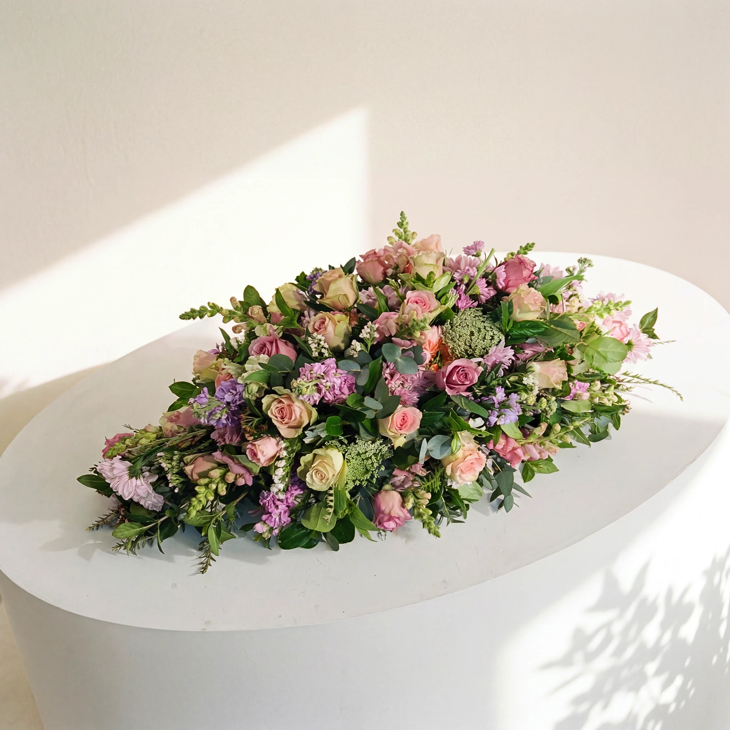 Forever Loved coffin spray of pink and lilac roses and stocks, displayed on a white plinth