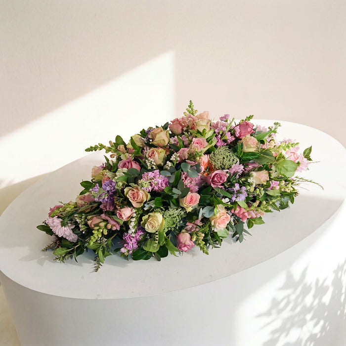 Forever Loved coffin spray of pink and lilac roses and stocks, displayed on a white plinth