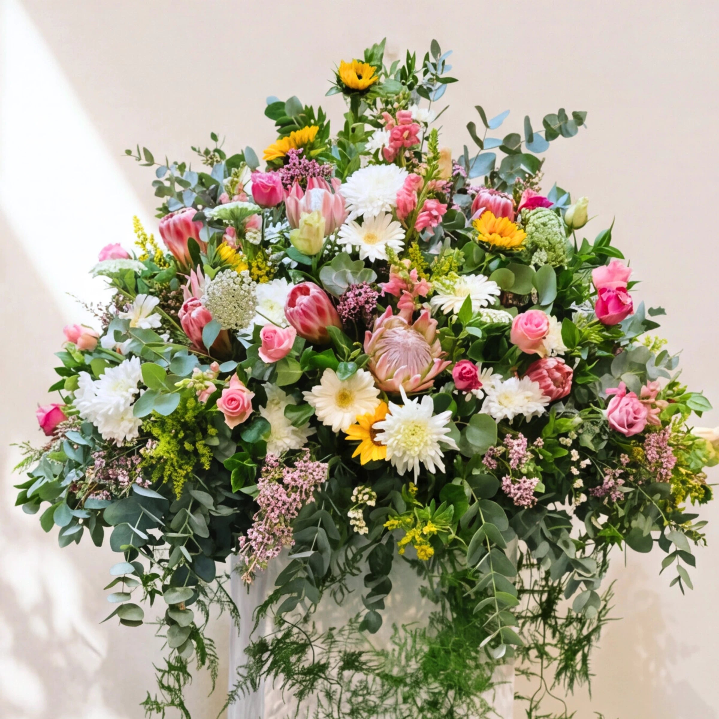 Large standing funeral tribute of pink proteas, roses, white chrysanthemums, sunflowers and eucalyptus.