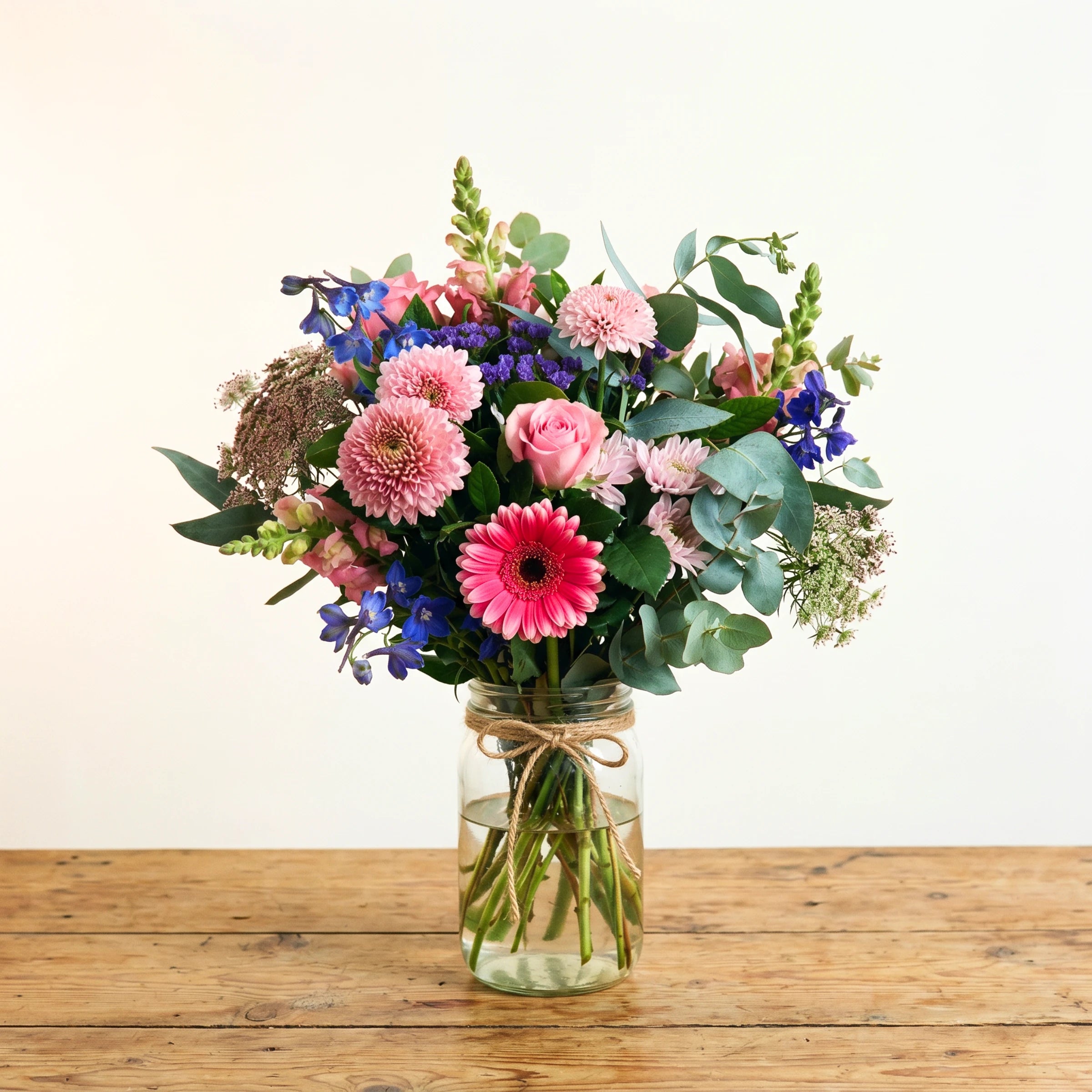 Glee in a Jar by Fabulous Flowers South Africa styled on a rustic wooden table, soft pastel pink, blue and purple flower arrangement with pink gerbera, chrysanthemums, delphinium and eucalyptus in a glass jar