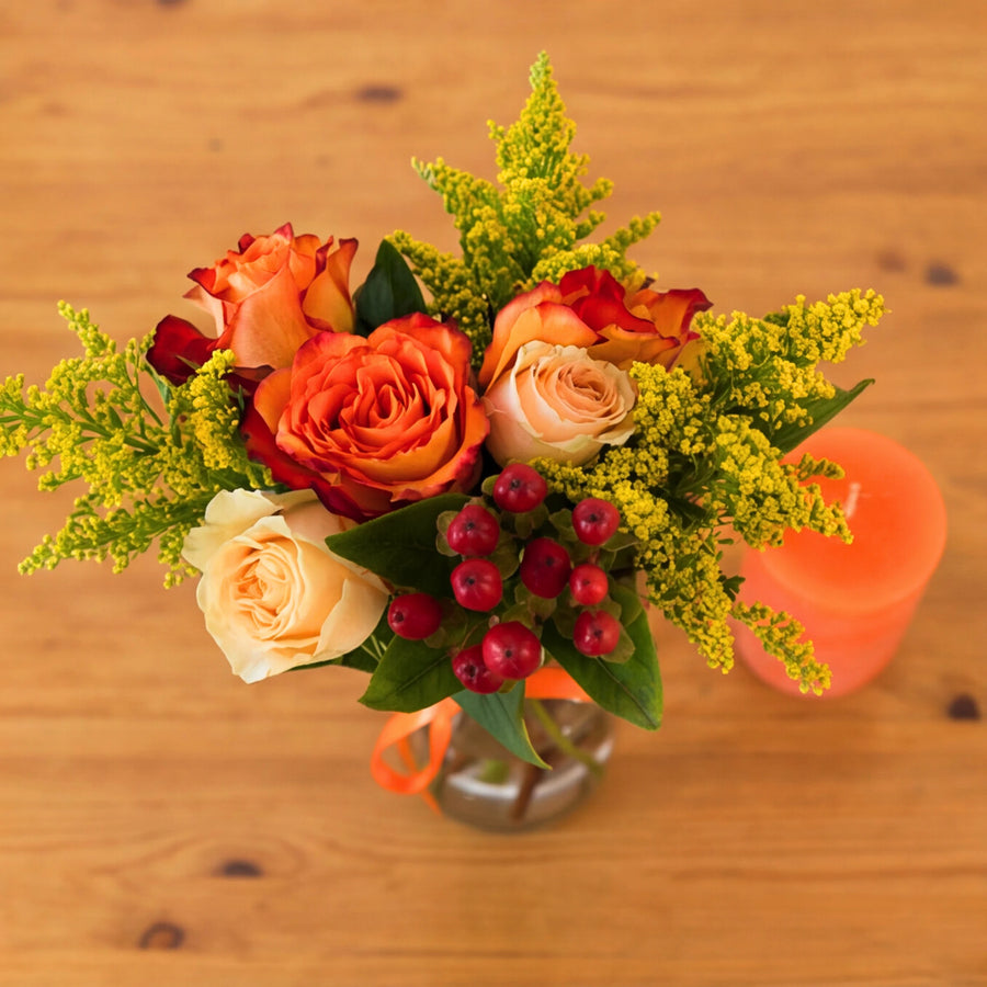 Top down view of orange and cream roses arranged in a vase with berries, solidago and an orange candle
