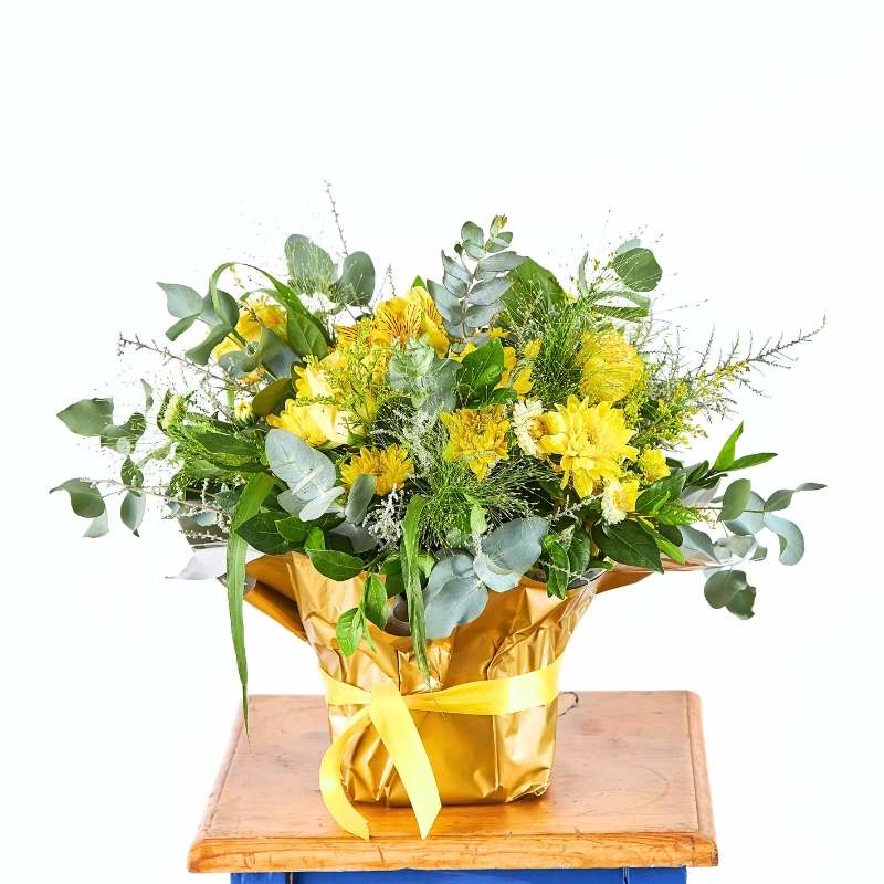 Arrangement of yellow flowers and greenery wrapped in gold paper on a wooden table with a white background