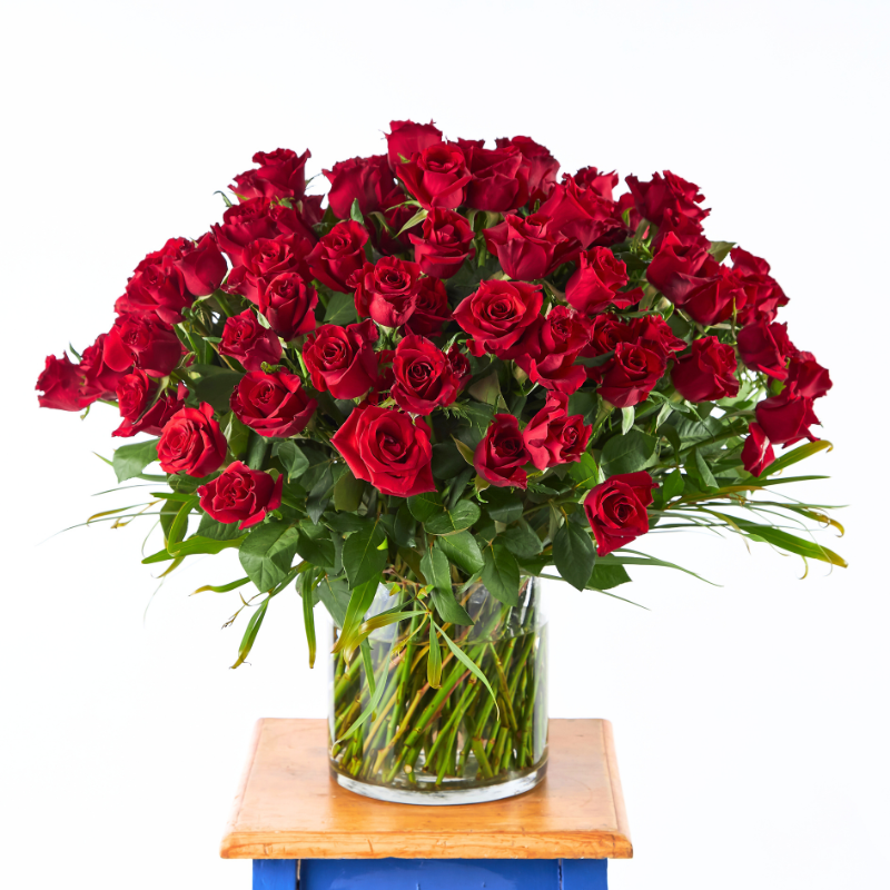 large ruby rose bouquet in a clear vase with green foliage, arranged on a blue and wooden table