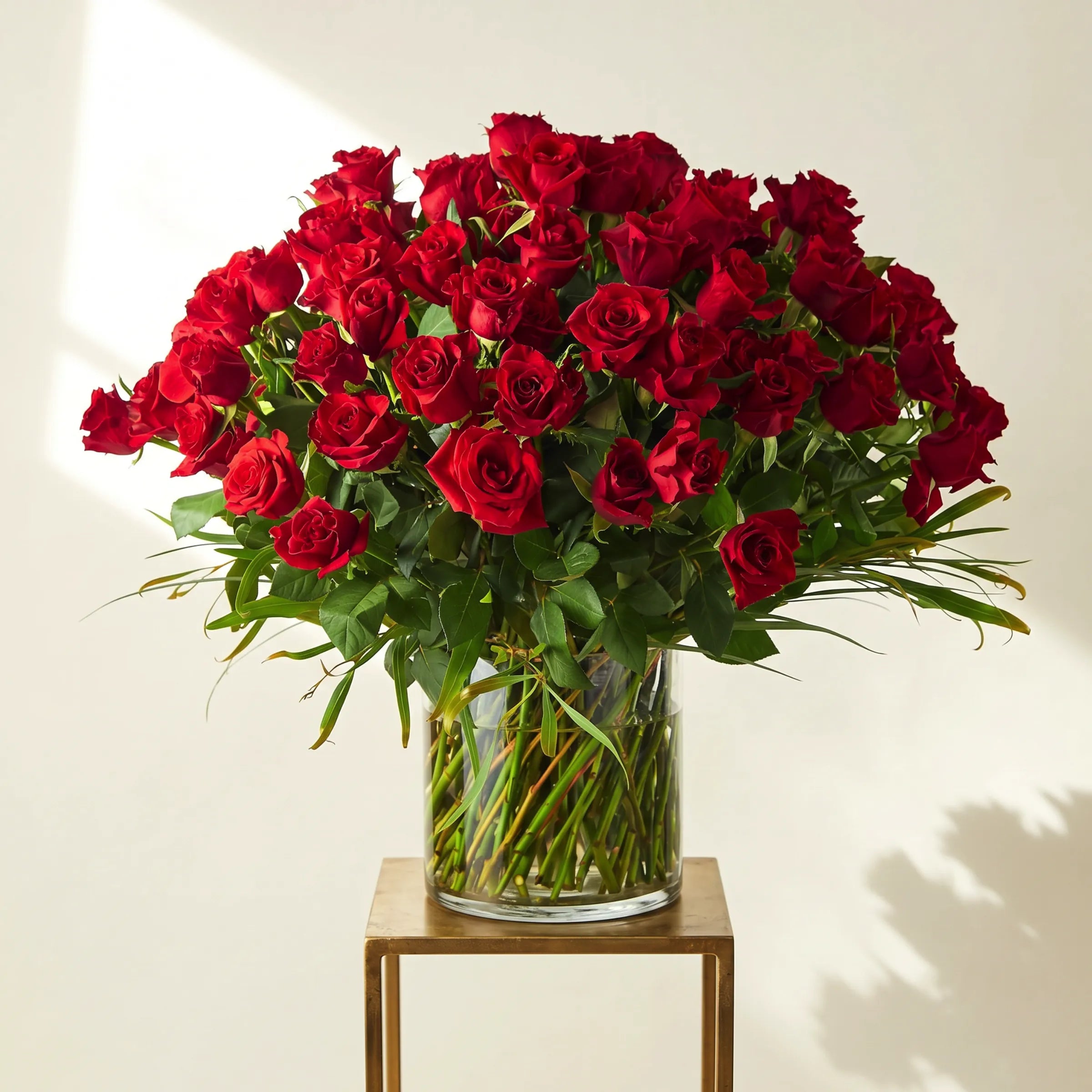 Golden Majesty arrangement of 100 luxury red roses in a clear glass cylinder vase displayed on a gold metal plinth in bright natural light, hand-arranged by Fabulous Flowers South Africa