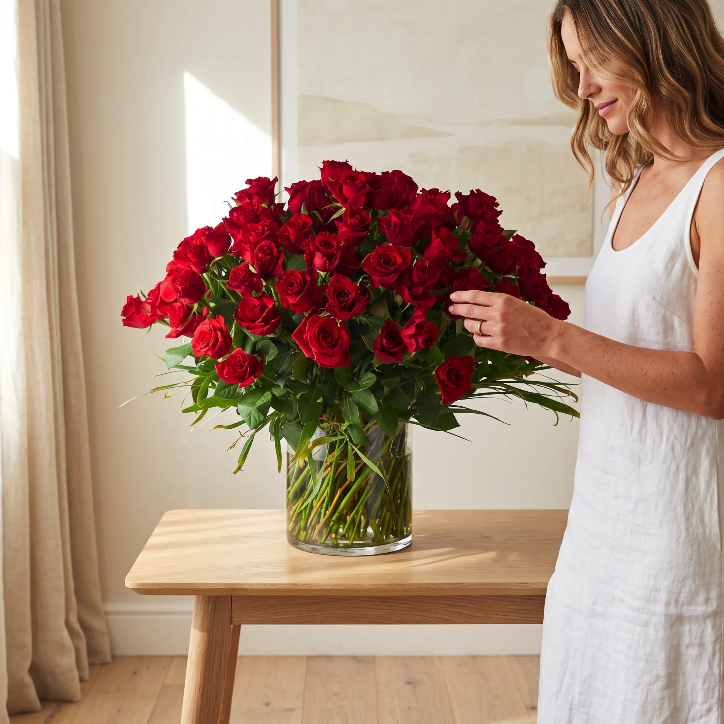 Woman in a white linen dress styling the Golden Majesty 100 red rose arrangement in a clear glass cylinder vase on a light oak console table, luxury red rose centrepiece by Fabulous Flowers South Africa anniversary and Valentine's gift
