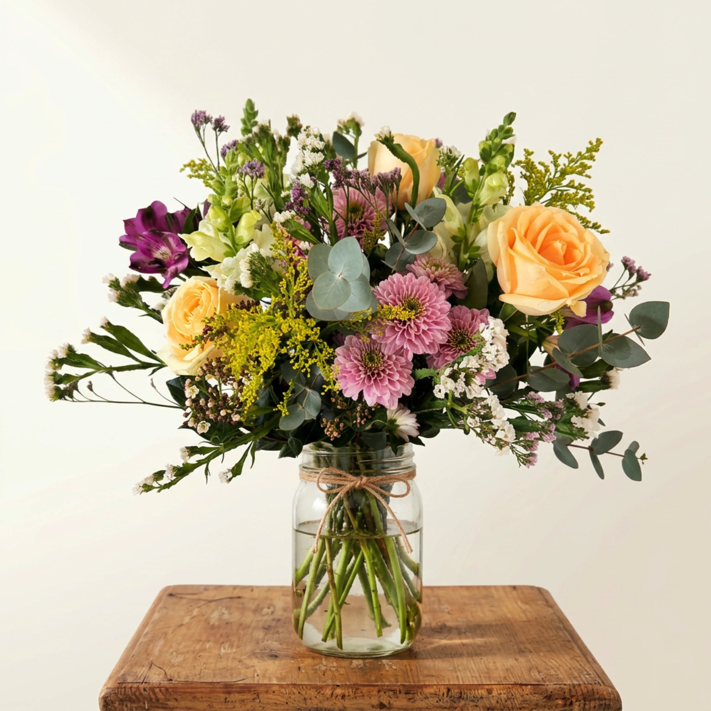 Grace in a Jar arrangement on a rustic wooden block, peach roses, lilac chrysanthemums, purple alstroemeria, white snapdragons, yellow solidago, lilac limonium and silver dollar eucalyptus in a recycled glass mason jar with twine bow, Fabulous Flowers Cape Town.