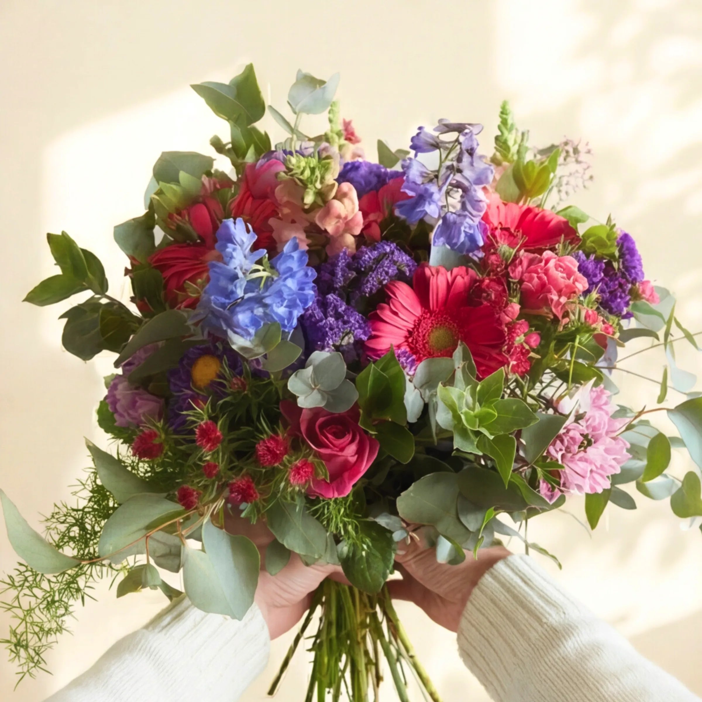 Hands holding the hand-tied Gratitude thank you bouquet of cerise, blue and purple blooms in warm Cape Town sunlight
