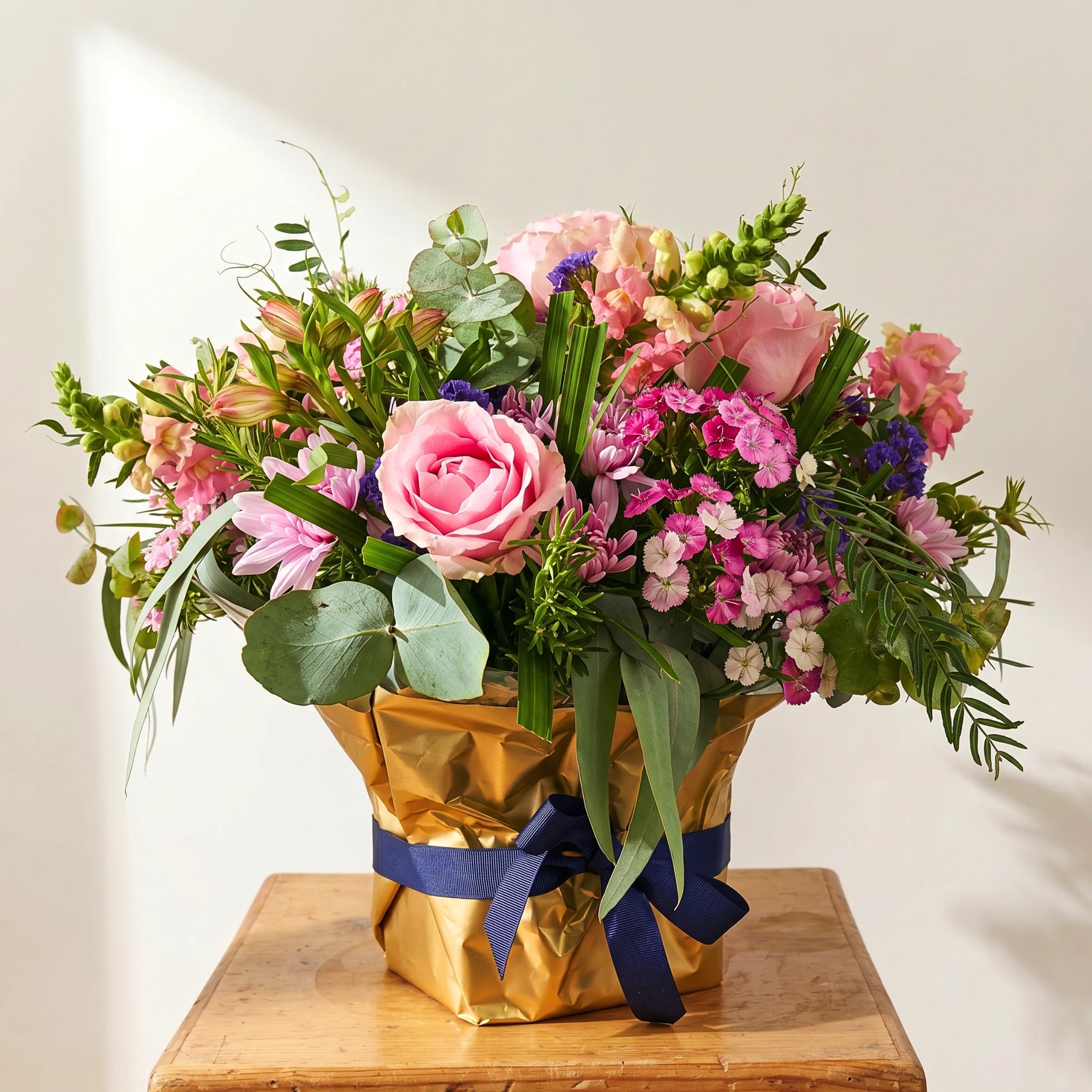 Harmony Flower Arrangement with pink roses, purple statice, pink alstroemeria, pink snapdragons, sweet william, lilac chrysanthemums and eucalyptus in a gold-wrapped container with navy ribbon bow on a wooden table, Fabulous Flowers Cape Town