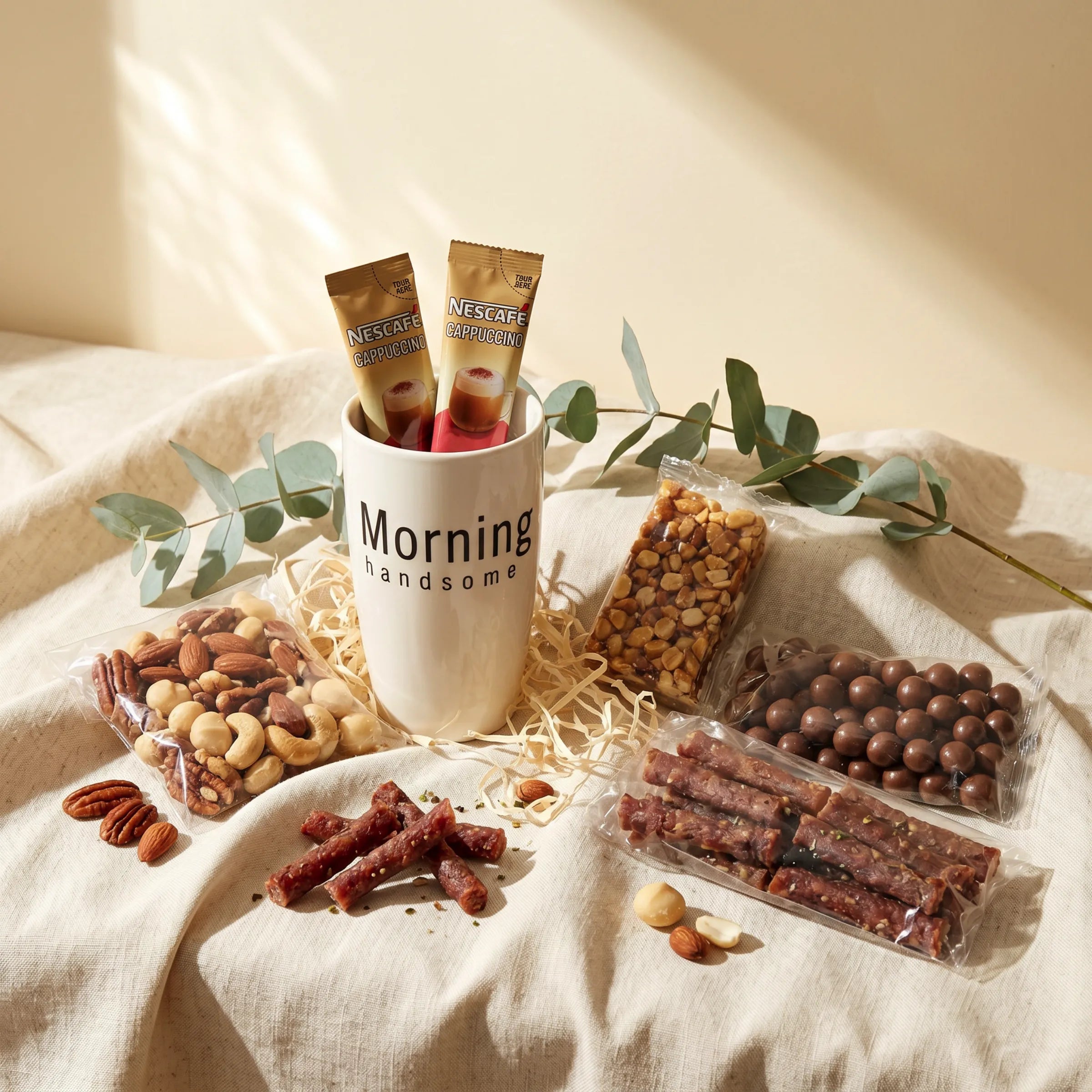 Hello Handsome Hamper flat-lay showing a white ceramic Morning Handsome mug with two Nescafé Cappuccino sachets, a bag of premium mixed nuts (pecans, almonds, cashews and macadamias), a peanut nougat bar, milk chocolate-coated balls and droëwors sticks on natural linen with eucalyptus styling, Fabulous Flowers South Africa gift hamper for him
