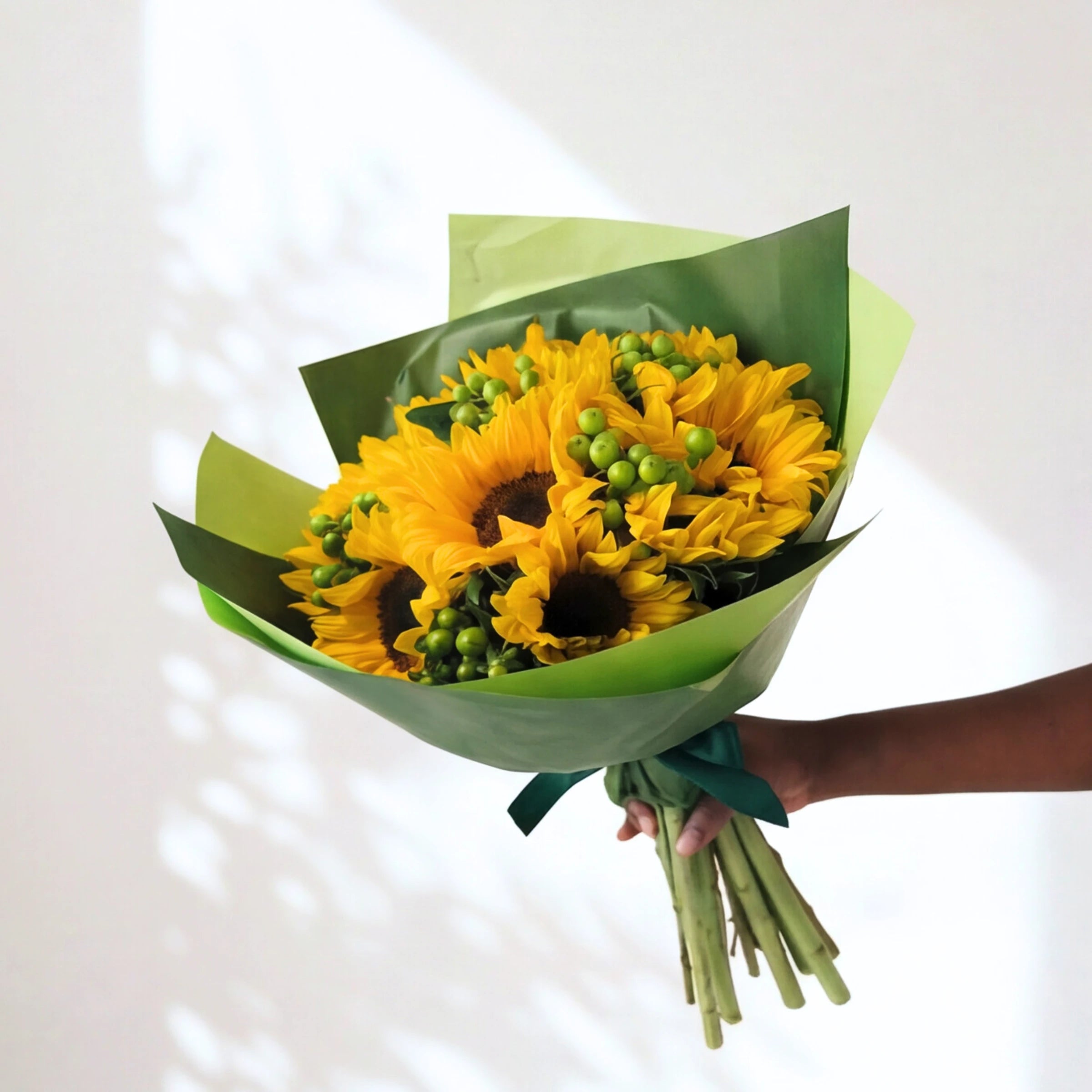 Here Comes the Sun bunch of sunflowers with green berries in green wrap, held against a sunlit wall
