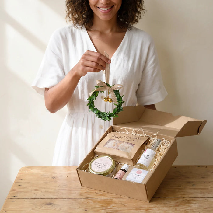 Woman in a white linen dress lifting a mini leaf wreath ornament out of an open kraft Christmas pamper box on a pine table, Fabulous Flowers South Africa