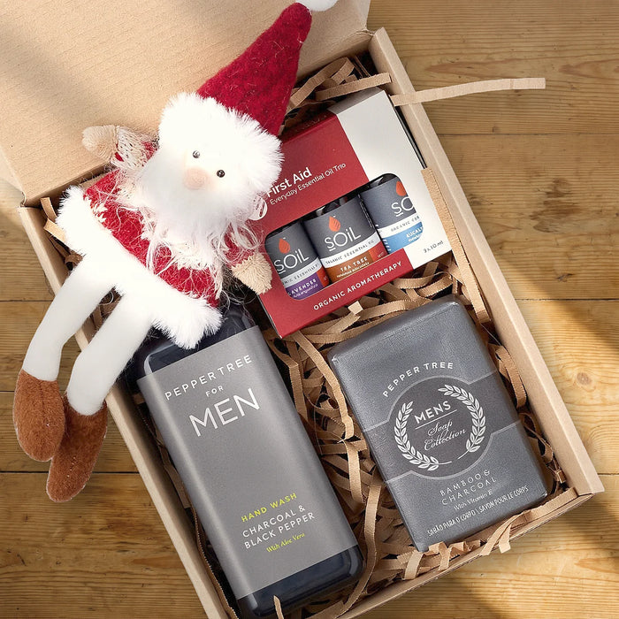 Open kraft gift box from above with a Peppertree for Men charcoal and black pepper hand wash, a Pepper Tree bamboo and charcoal soap, a SOiL essential oil trio and a plush Santa on a sunlit pine table, Fabulous Flowers South Africa